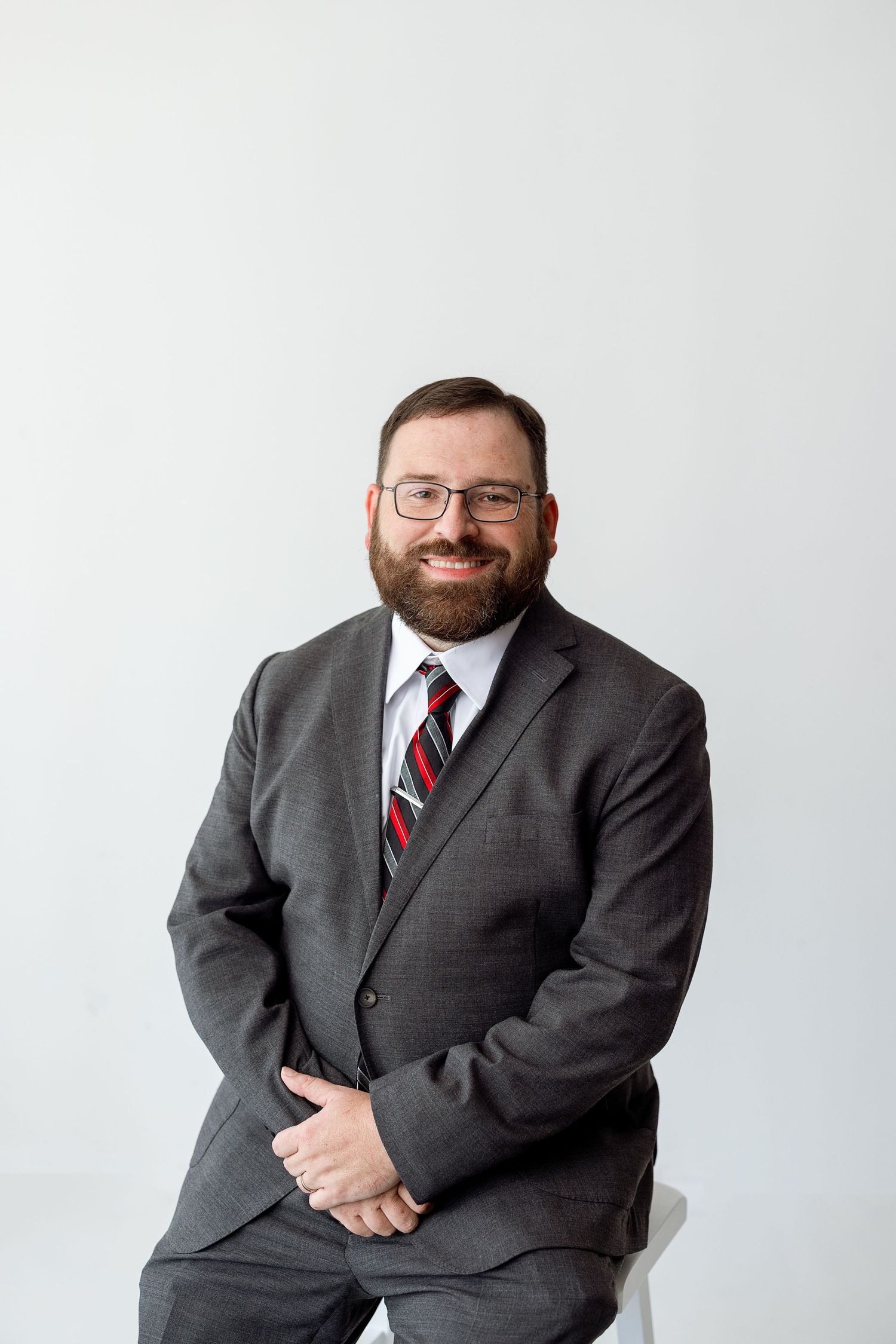 Professional studio portrait of someone in a dark gray suit with a red striped tie sitting and posing against a light background.