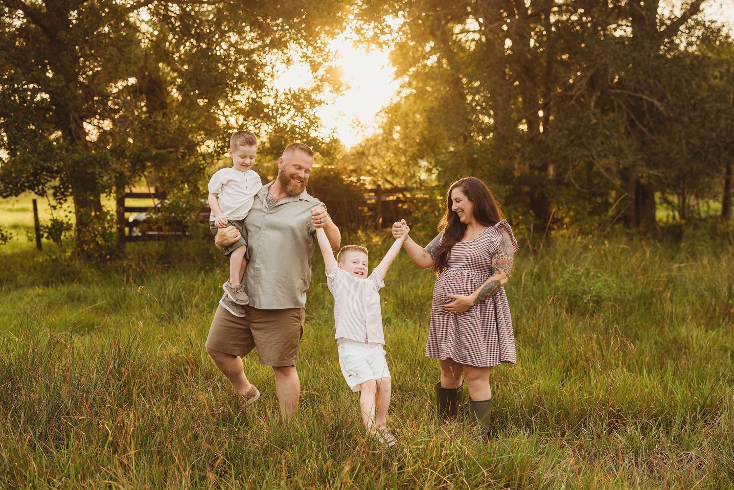 Pregnant woman and man with two young children playing in a sunlit green field during a golden hour family maternity photo session.