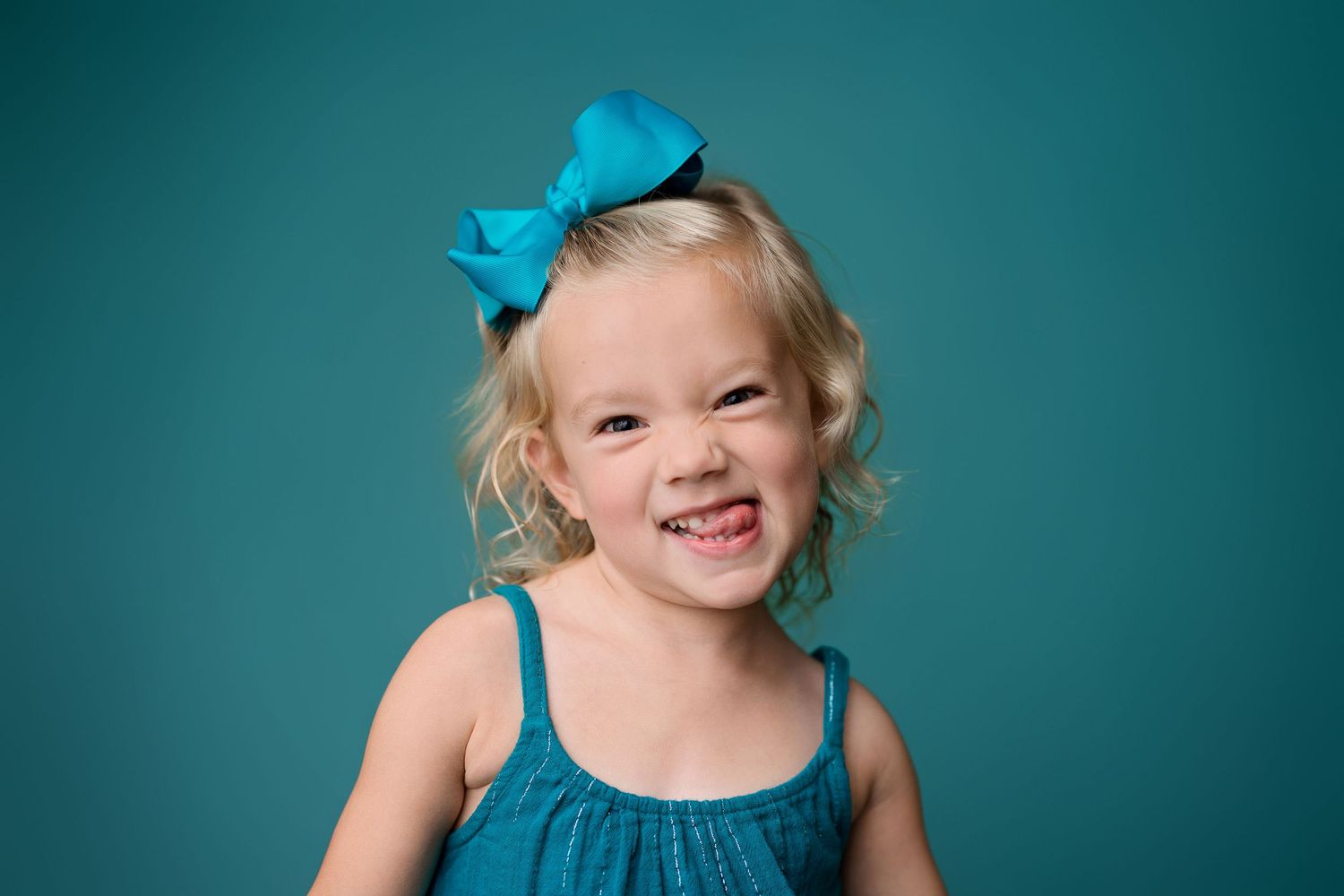 A toddler wearing a teal blue bow and dress poses and laughs joyfully against a turquoise studio backdrop.