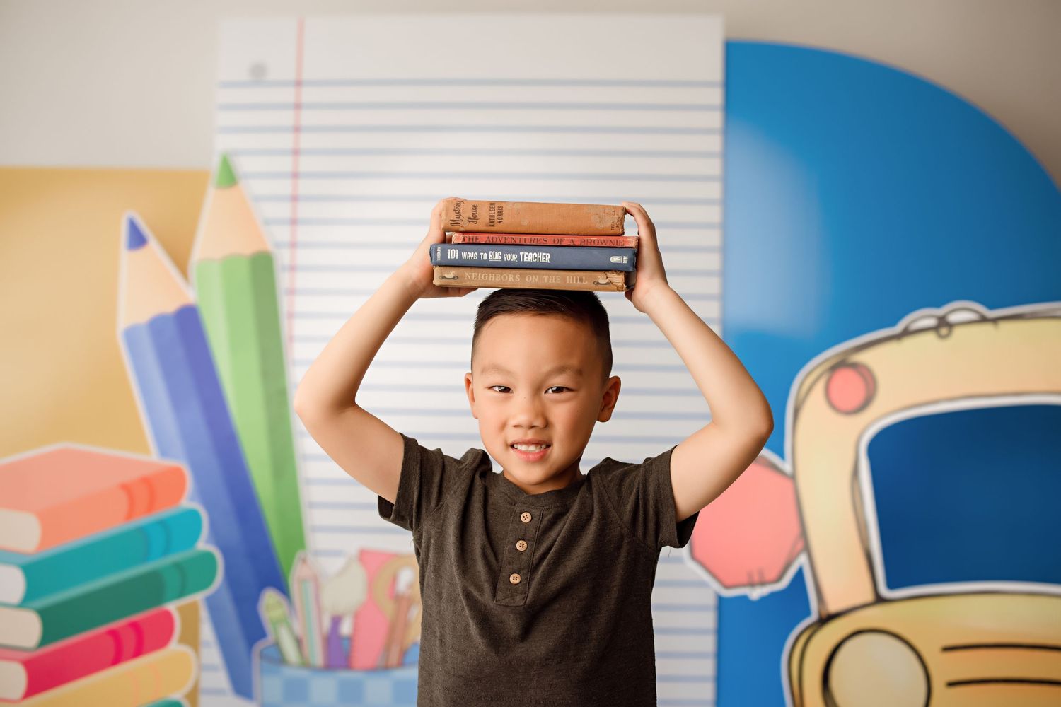 Child holding books above head poses in front of school-themed backdrop.