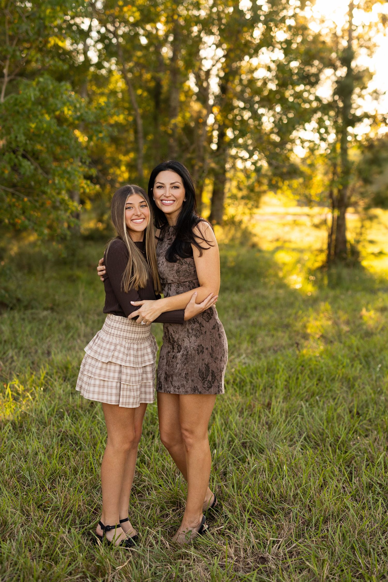 Two friends embrace while standing in a grassy field at golden hour with sunlit trees in the background.
