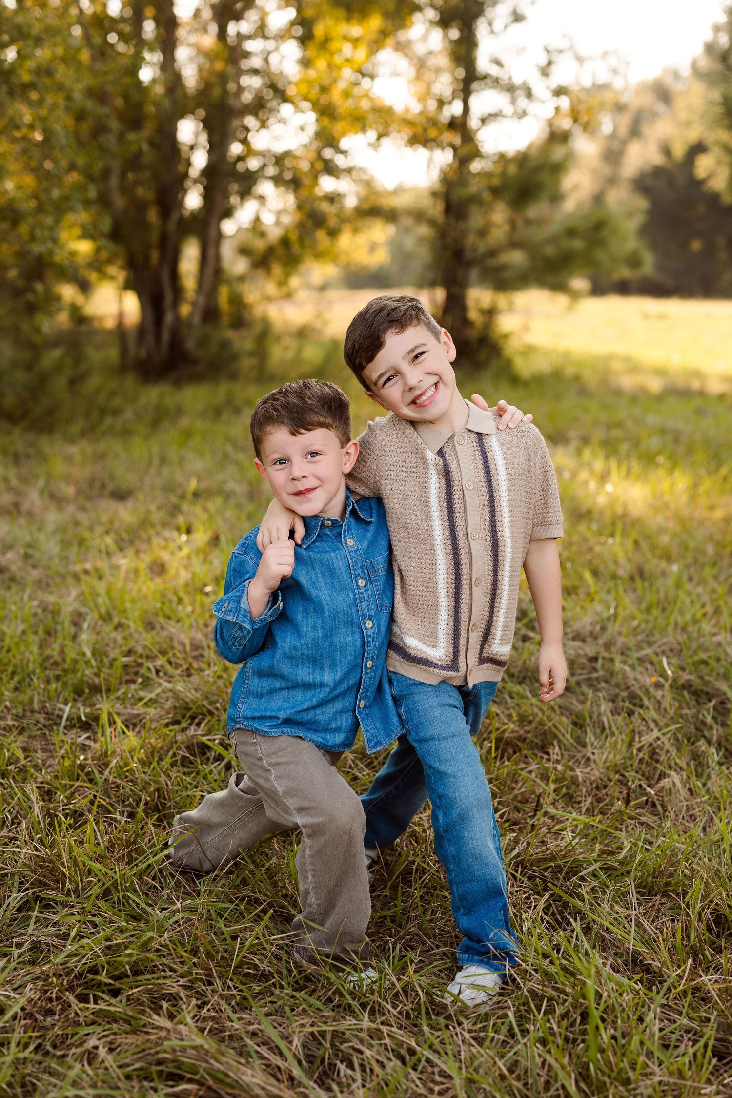 Two smiling young boys in casual clothes pose with arms around each other in a sunlit grassy field with trees behind them.