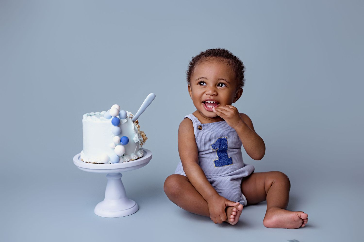 A cake smash photo session shows a baby in a light colored outfit next to a white birthday cake with blue decorations.