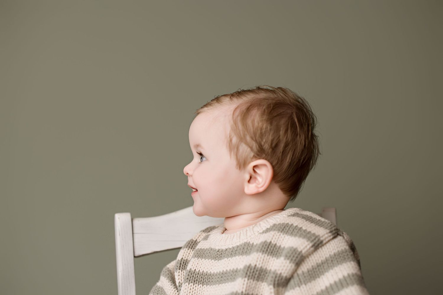 Baby in white rocking chair looks to the side while wearing cozy neutral-toned clothing against beige background.