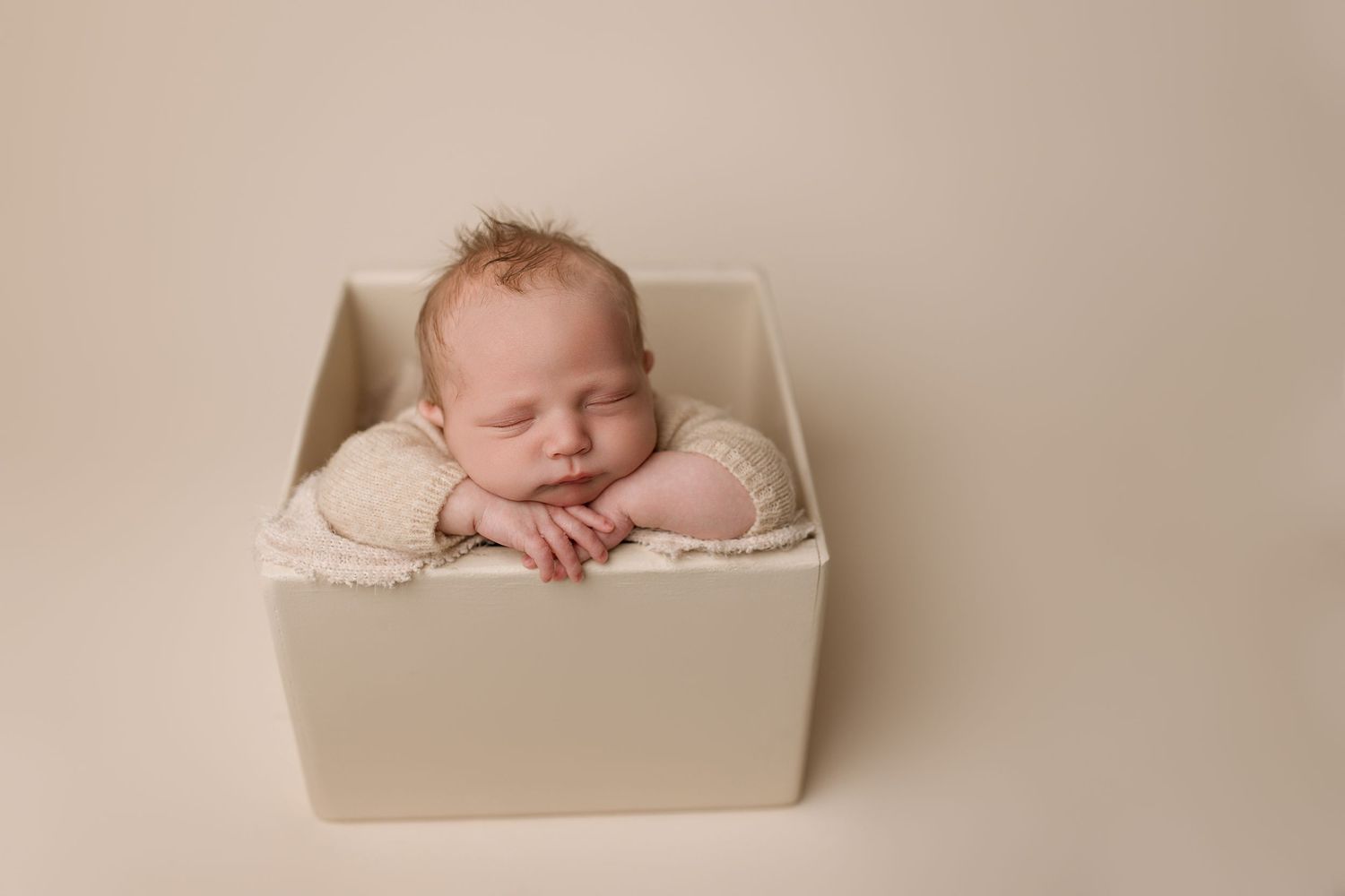 A sleeping newborn baby lies peacefully in a cream-colored photography prop during a professional photo session.