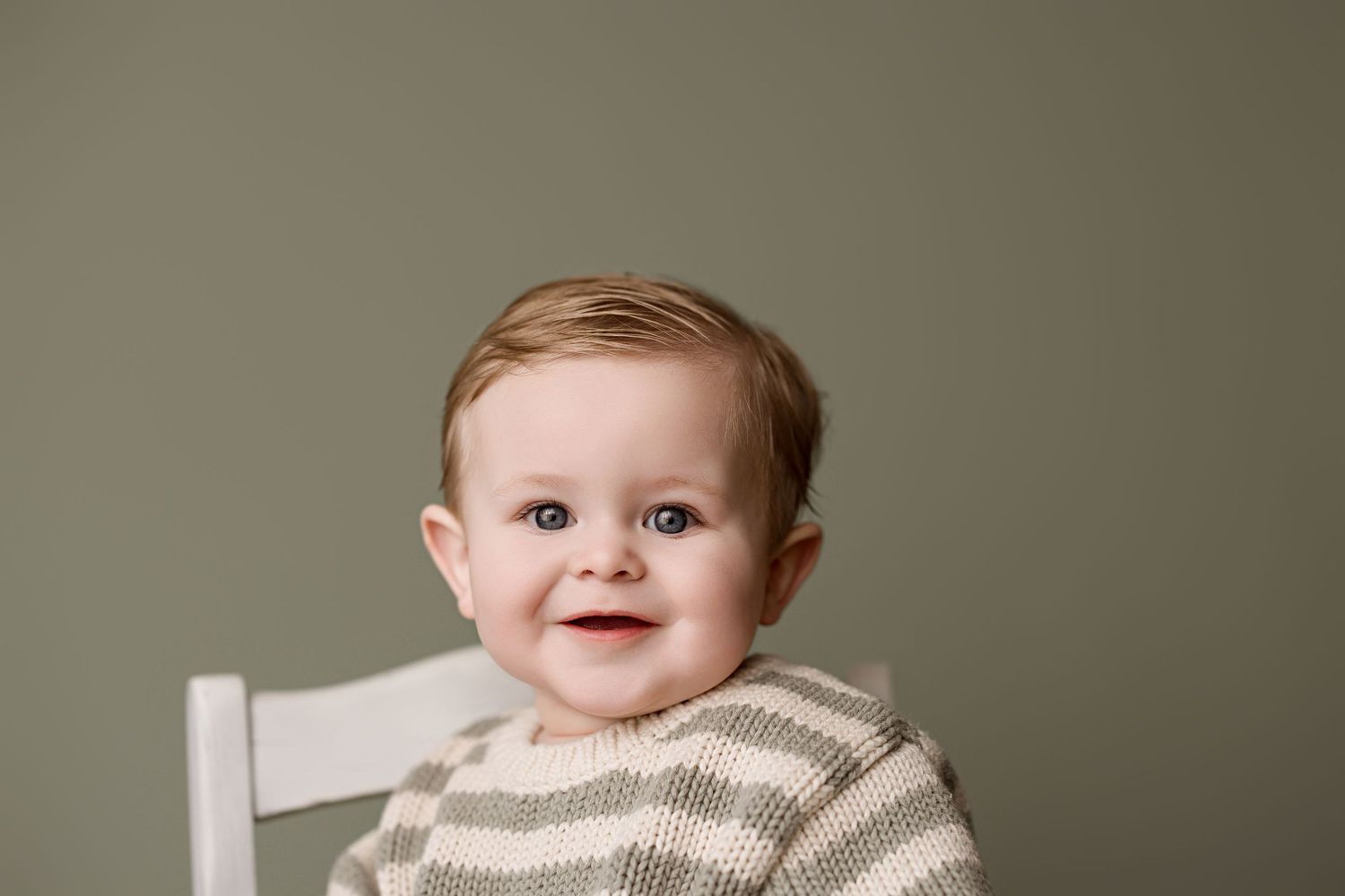 A smiling baby wearing a gray and white striped sweater sits in a white chair against a neutral background.