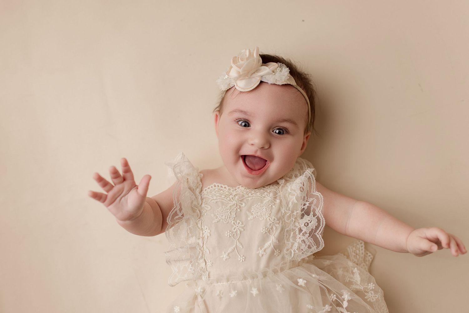 A baby wearing a white lacy christening gown and headband smiles while laying on a beige backdrop.