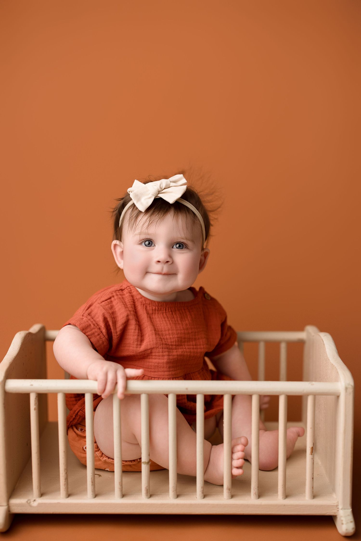 Baby wearing rust-colored outfit sits in wooden doll crib against orange background.
