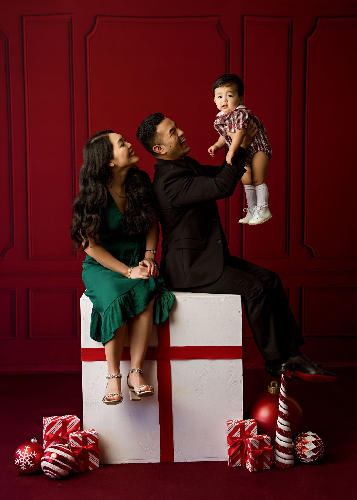 A family poses on a white gift box prop against a red wall with Christmas decorations.
