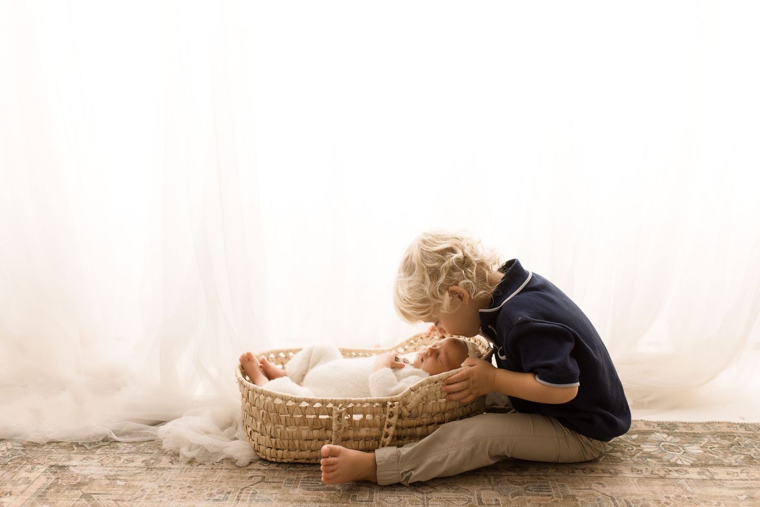 Siblings lying on white blanket near wicker basket in natural light setting.