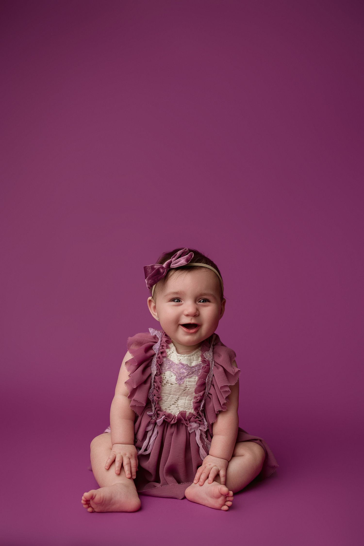 Baby wearing dusty rose dress sits and smiles on purple backdrop in studio portrait series.