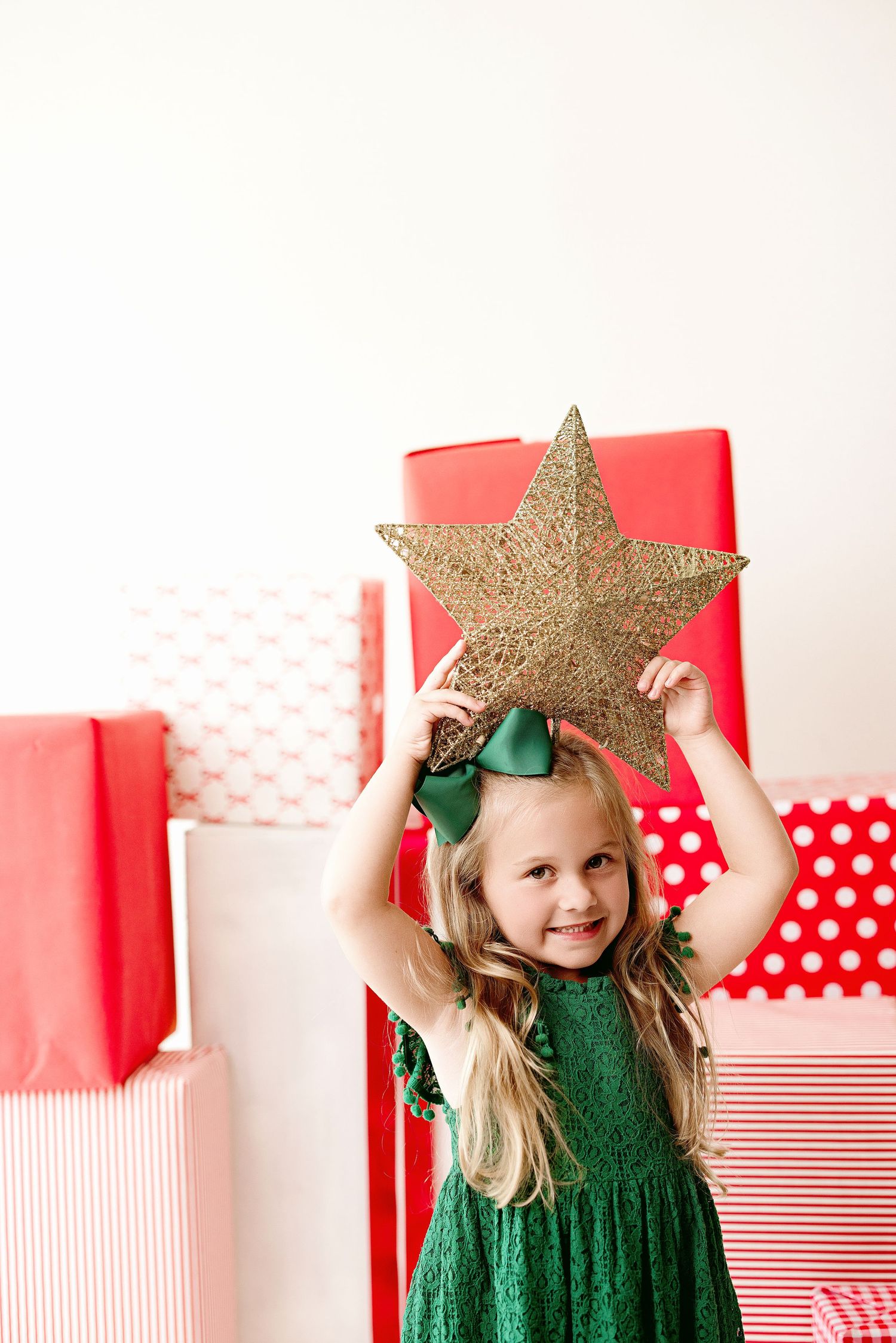 Young girl in green dress holds gold star decoration against backdrop of red and white Christmas presents.