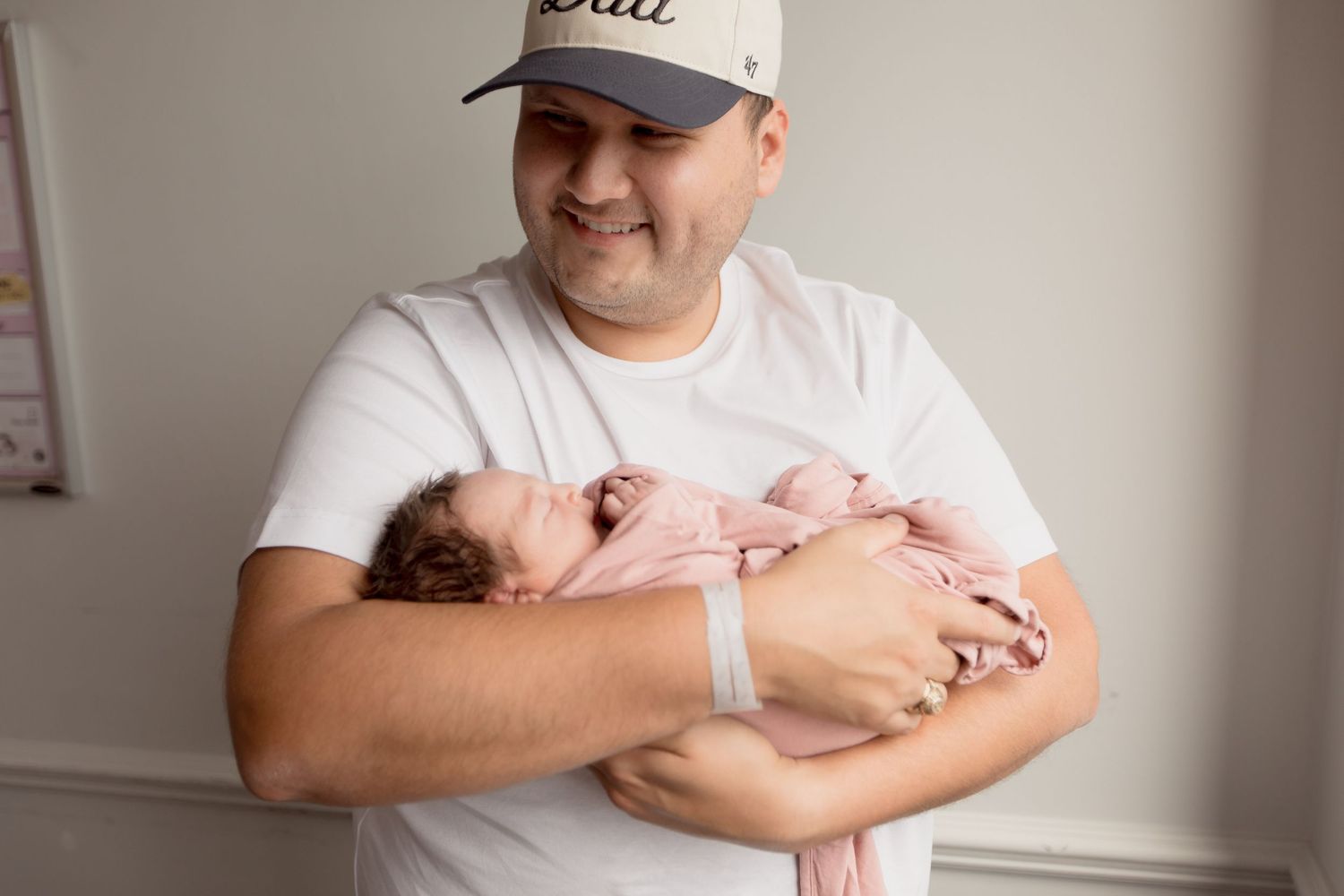 A person wearing a white baseball cap and t-shirt holds a sleeping newborn baby wrapped in pink.