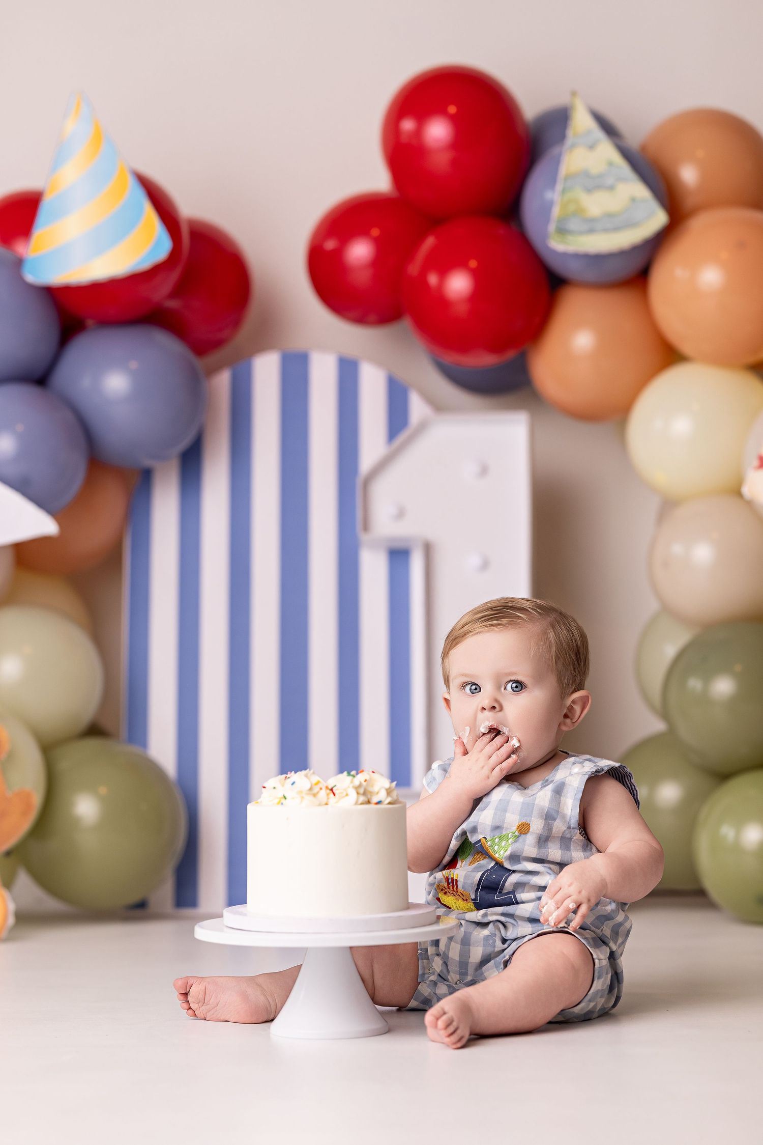 Baby in striped romper poses with cake and colorful balloon garland during first birthday cake smash photo session.
