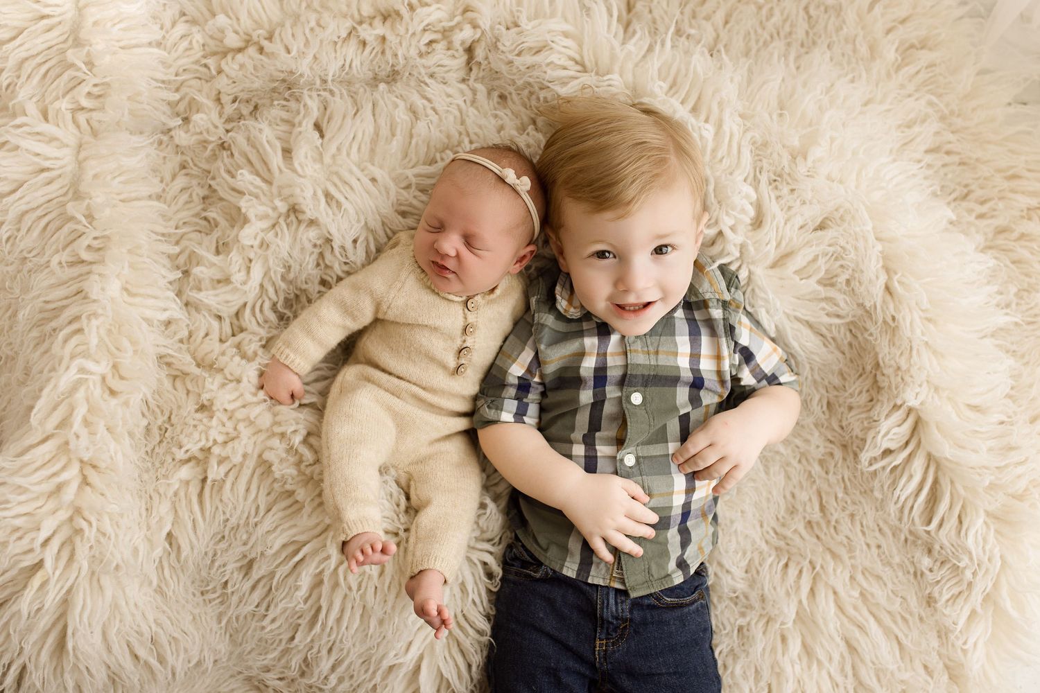 Two siblings lying on fluffy white rug with older child in plaid shirt hugging newborn baby.