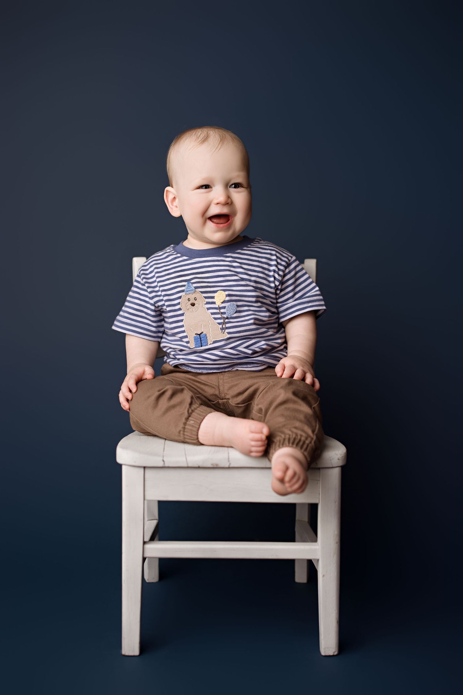 A baby in a striped shirt and brown pants sits on a small white bench against a dark blue background.