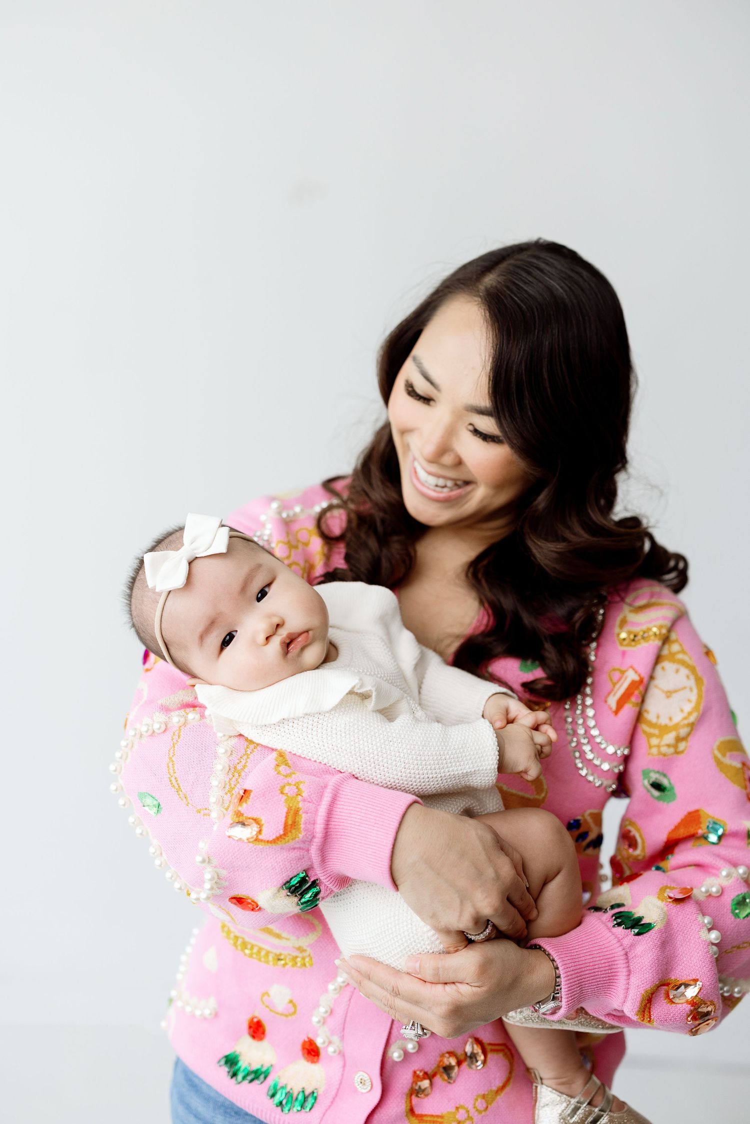 Tender moment as mother in pink floral kimono holds smiling baby in white dress against white backdrop.