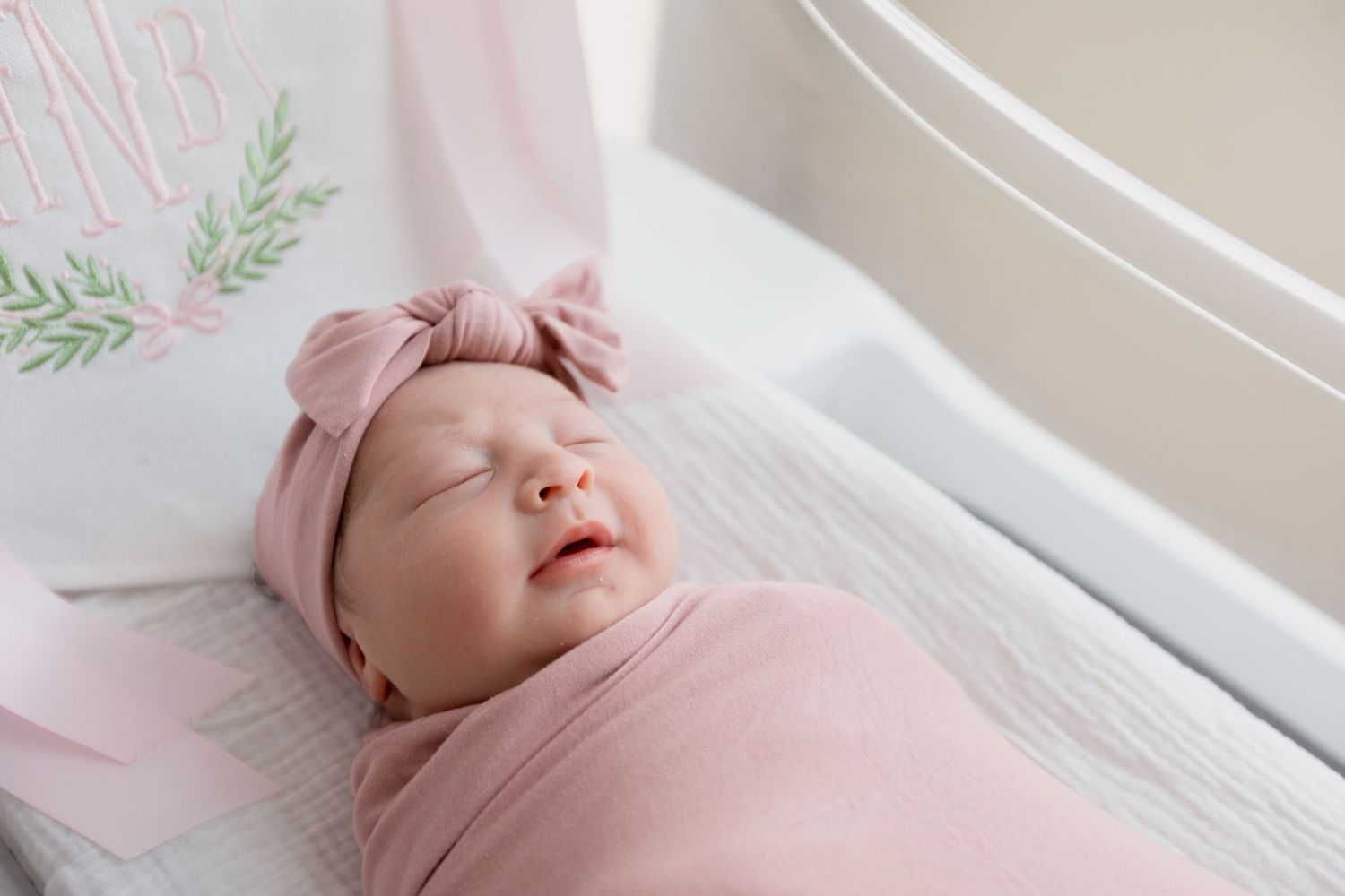 A sleeping baby lies peacefully in a bright white bassinet near a sunlit window during Fresh 48 session in Beaumont hospital.