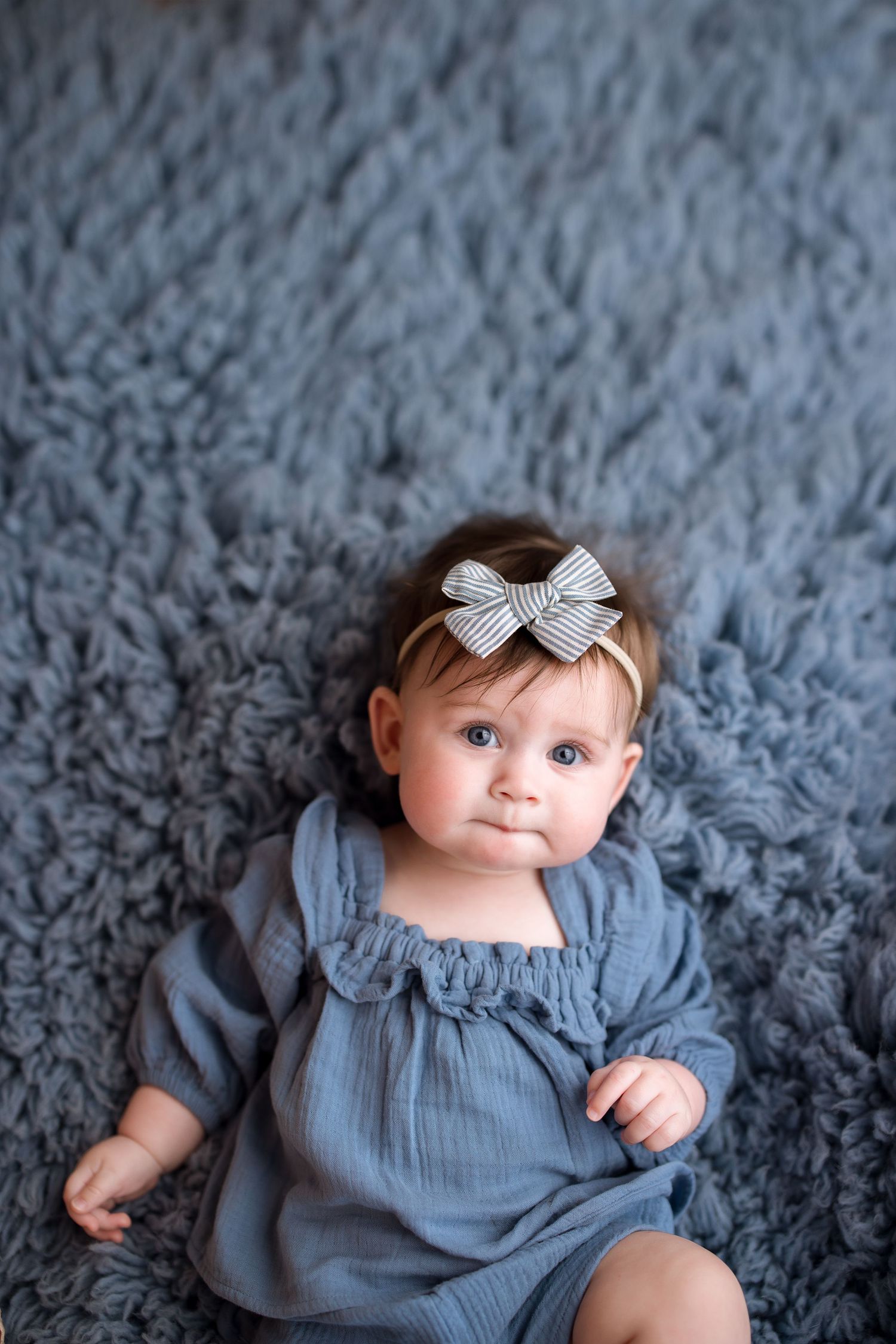 Baby in blue dress lying on gray textured blanket with white bow headband.