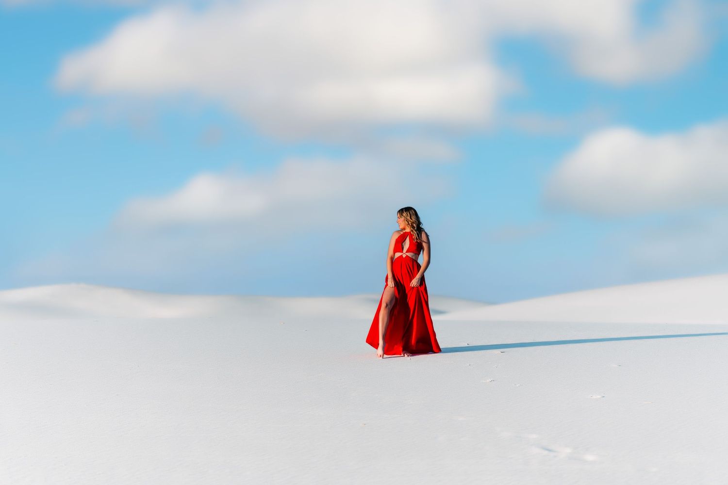 A person in a red dress stands alone against a stark white snowy landscape beneath a bright blue sky.