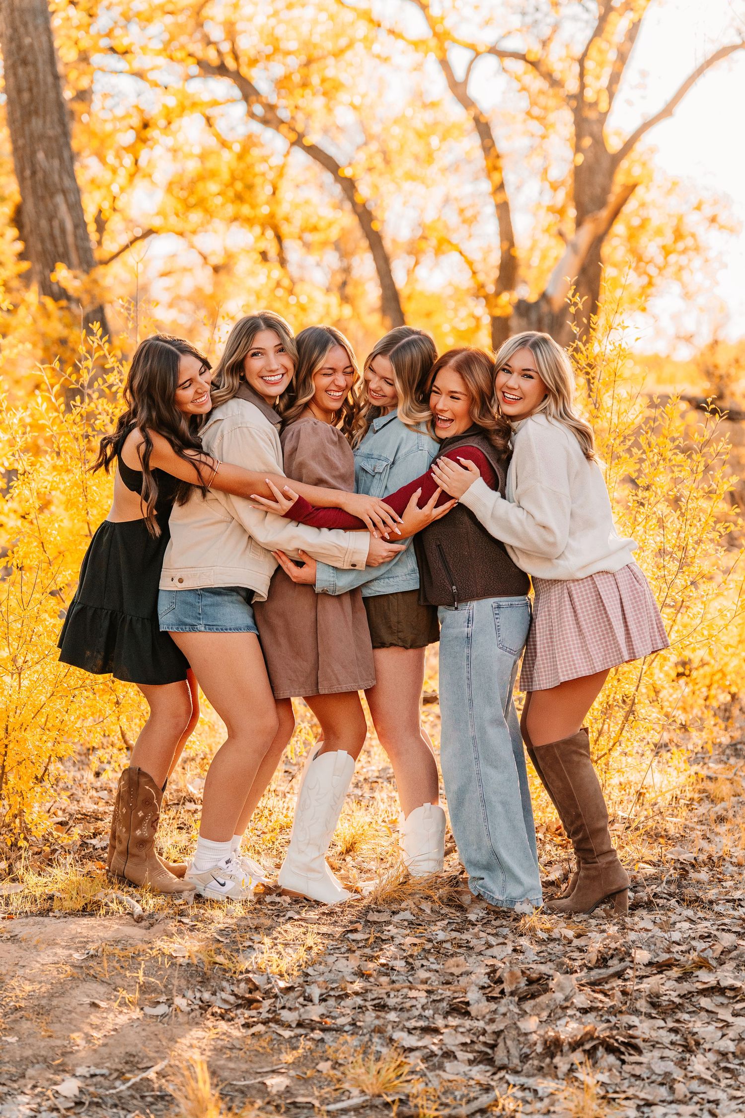 Group photo of friends laughing and embracing on fallen tree with golden autumn trees in background.