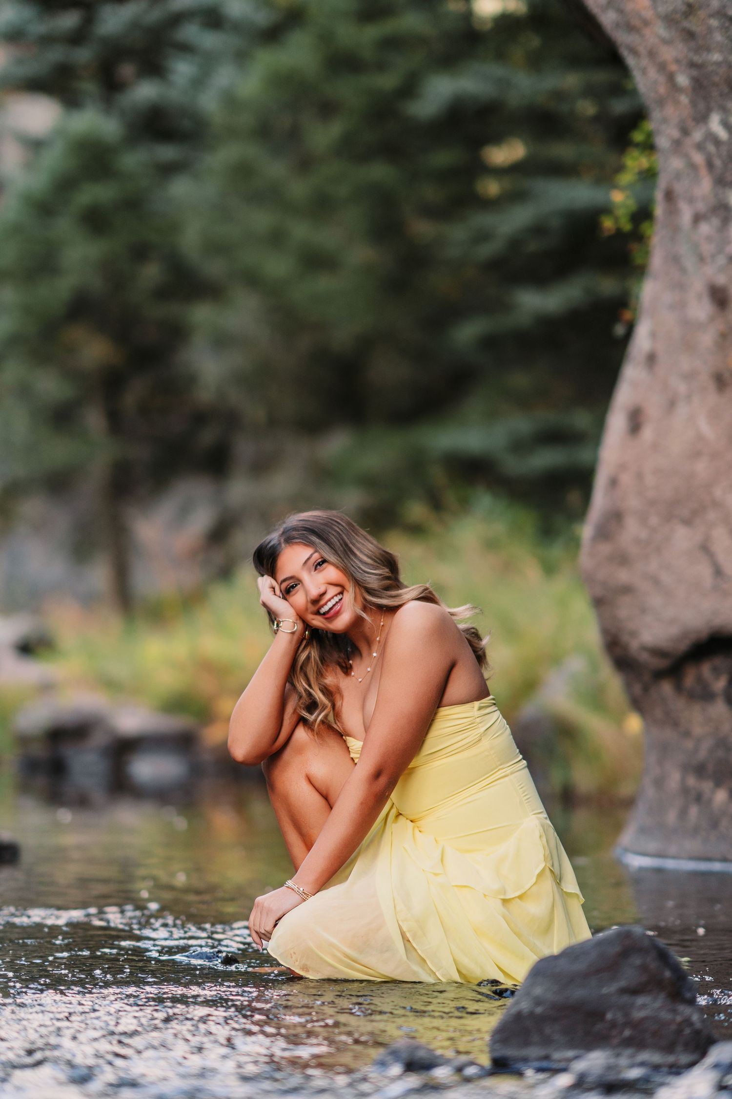 A person in a yellow dress poses playfully by a stream among rocks and trees.