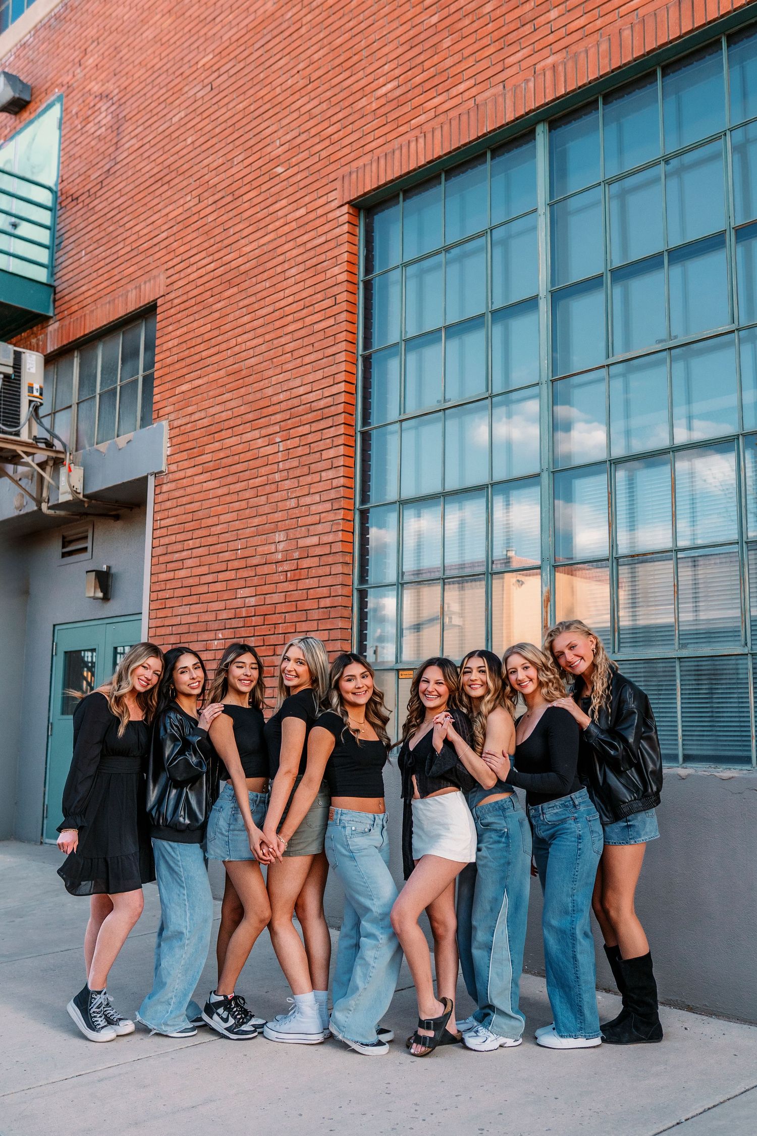 Group poses playfully against brick wall wearing matching denim and black jackets.