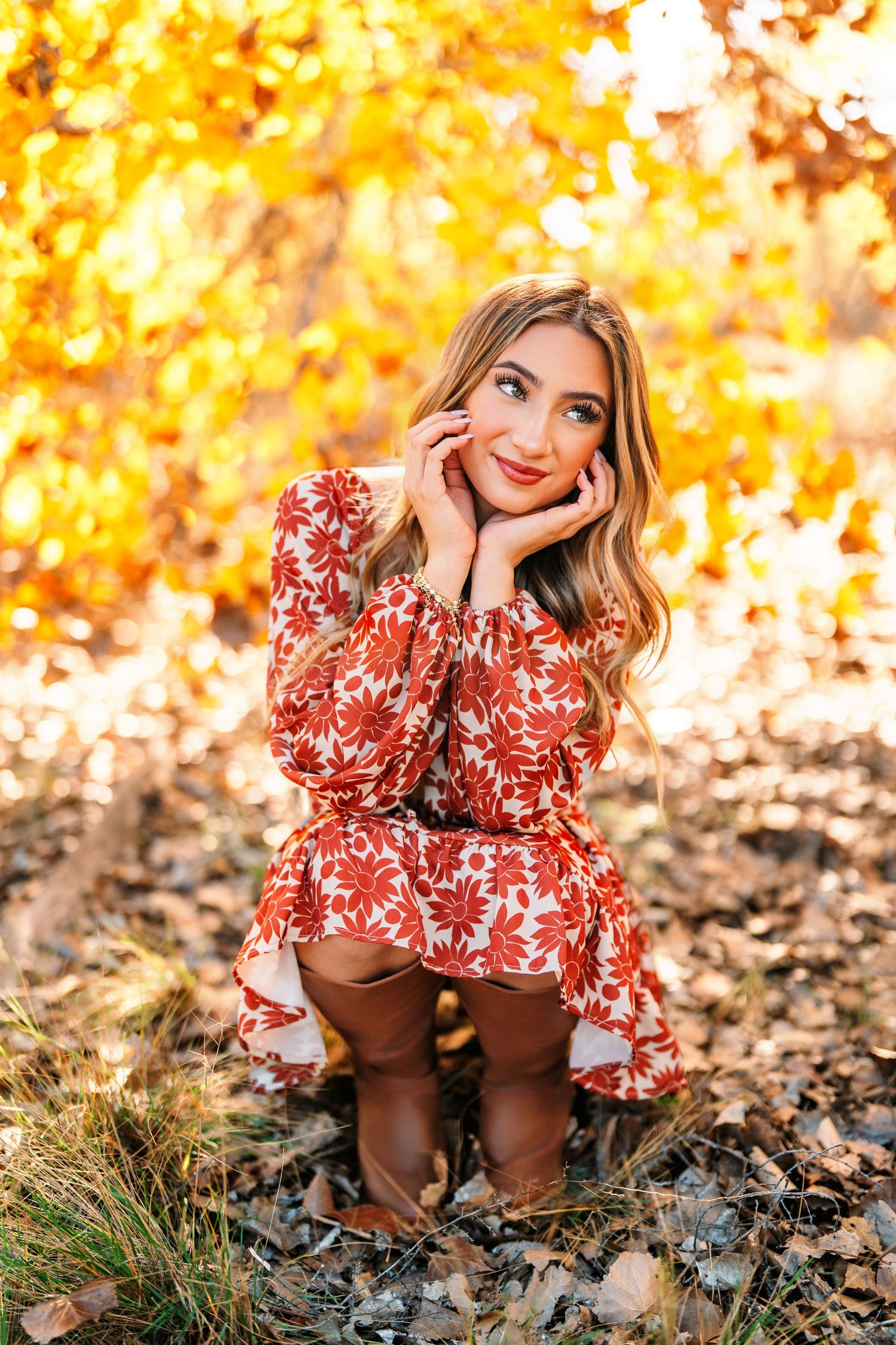 Woman in red floral dress crouches among autumn leaves with golden bokeh background.