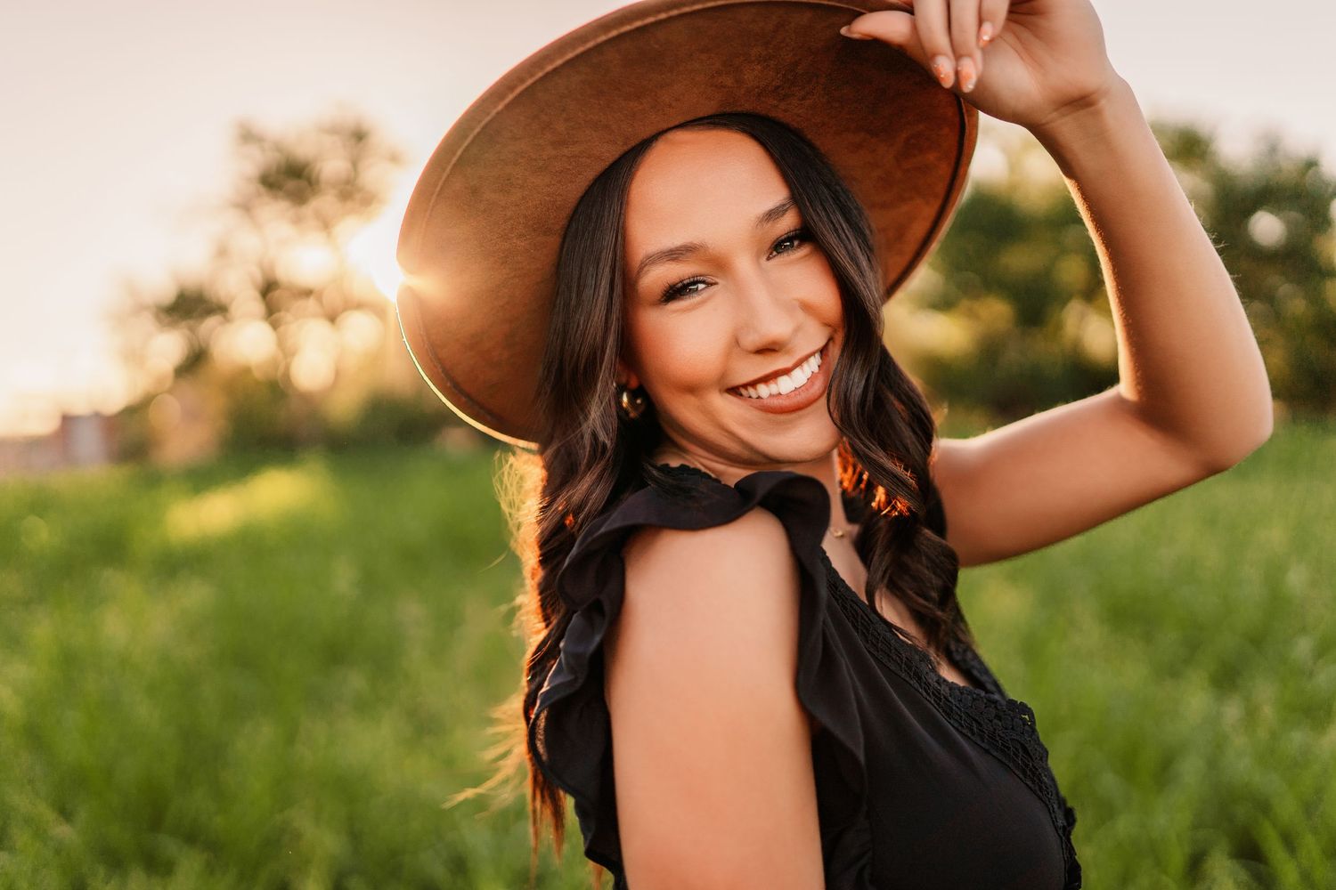 Woman in black dress and brown hat poses in a sunny field during golden hour.