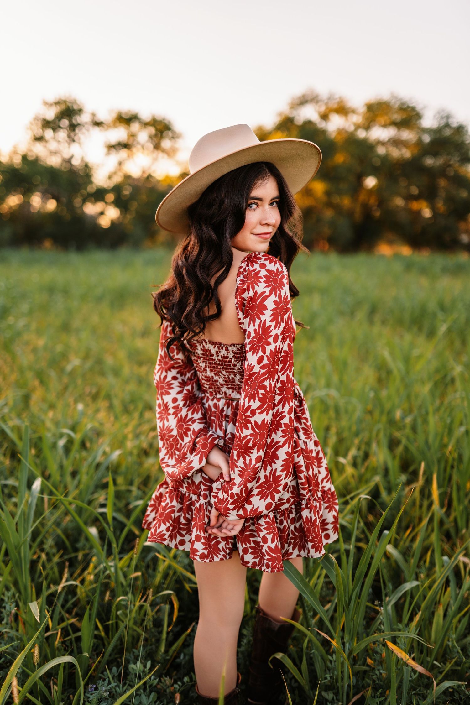 Sunset portraits in flowing red floral dress and hat among tall grass.