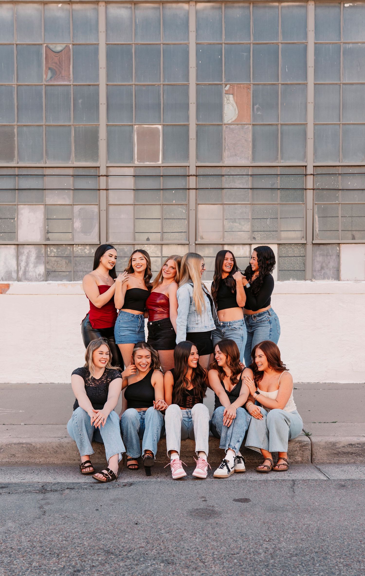 Group sitting casually against industrial wall with windows wearing denim and casual attire.