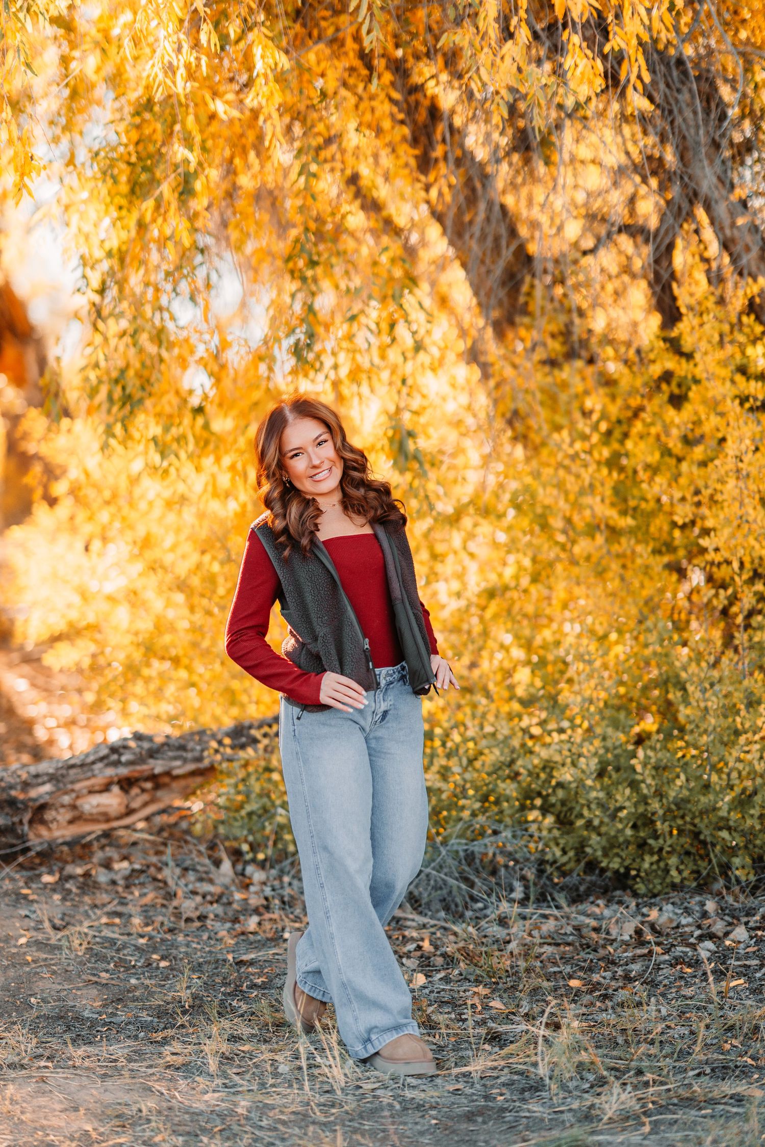 Person in casual outfit sits on ground with golden fall foliage in background during sunset photo session.