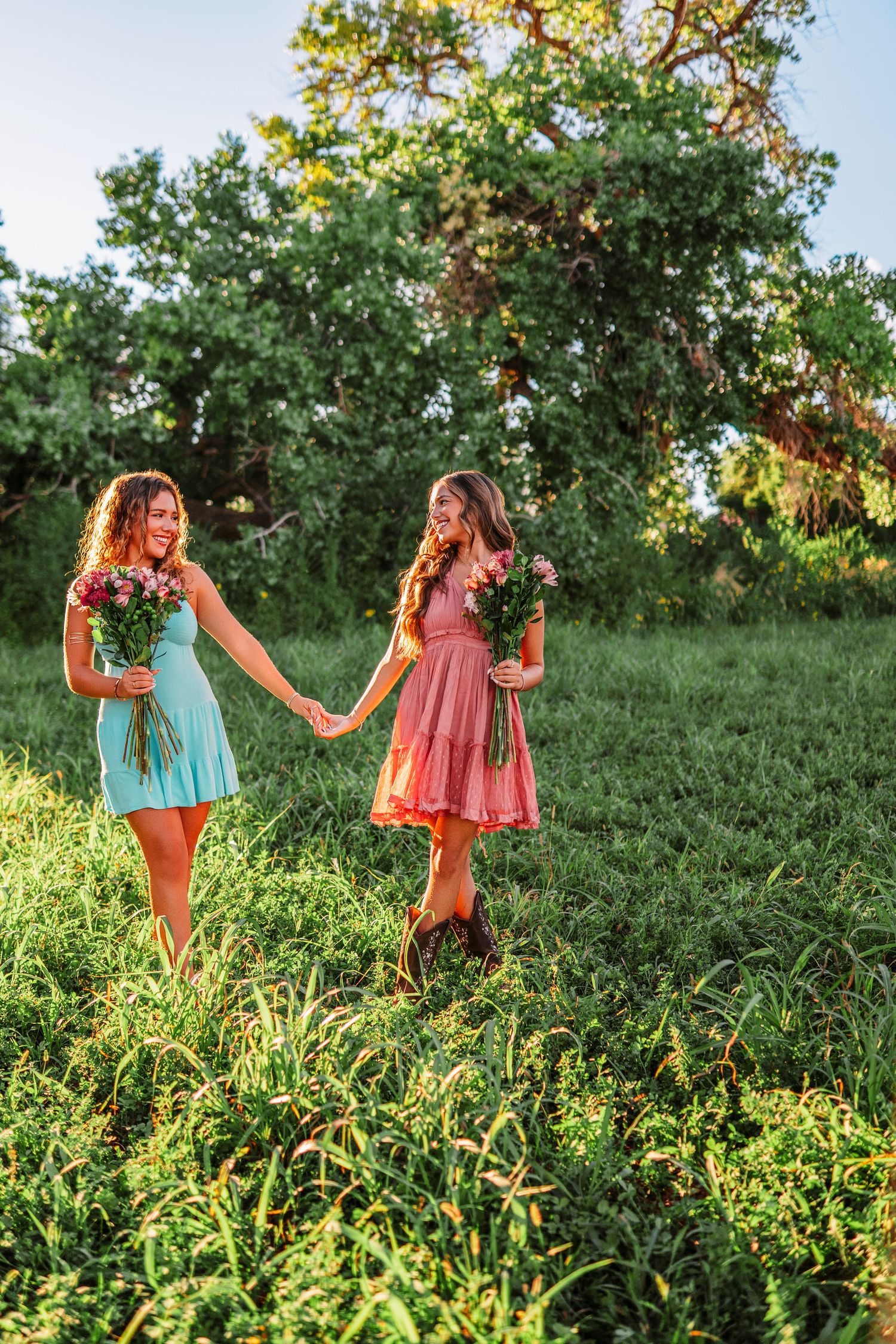 Friends in colorful summer dresses walking through a sunlit field with trees.
