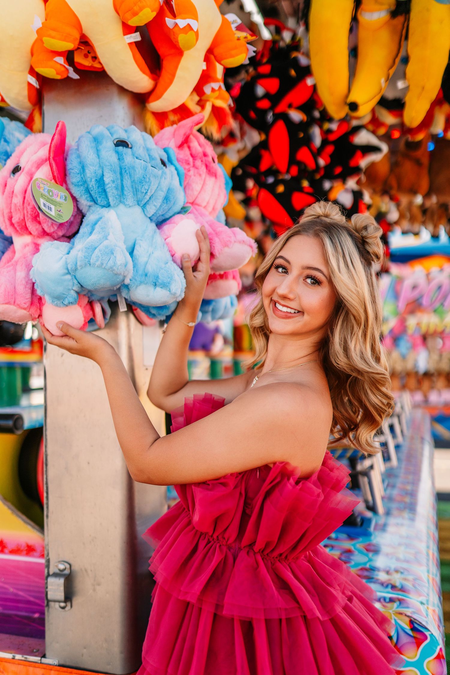 Playful carnival photoshoot with stuffed animal prizes and bright pink dress against vibrant game booth backdrop.