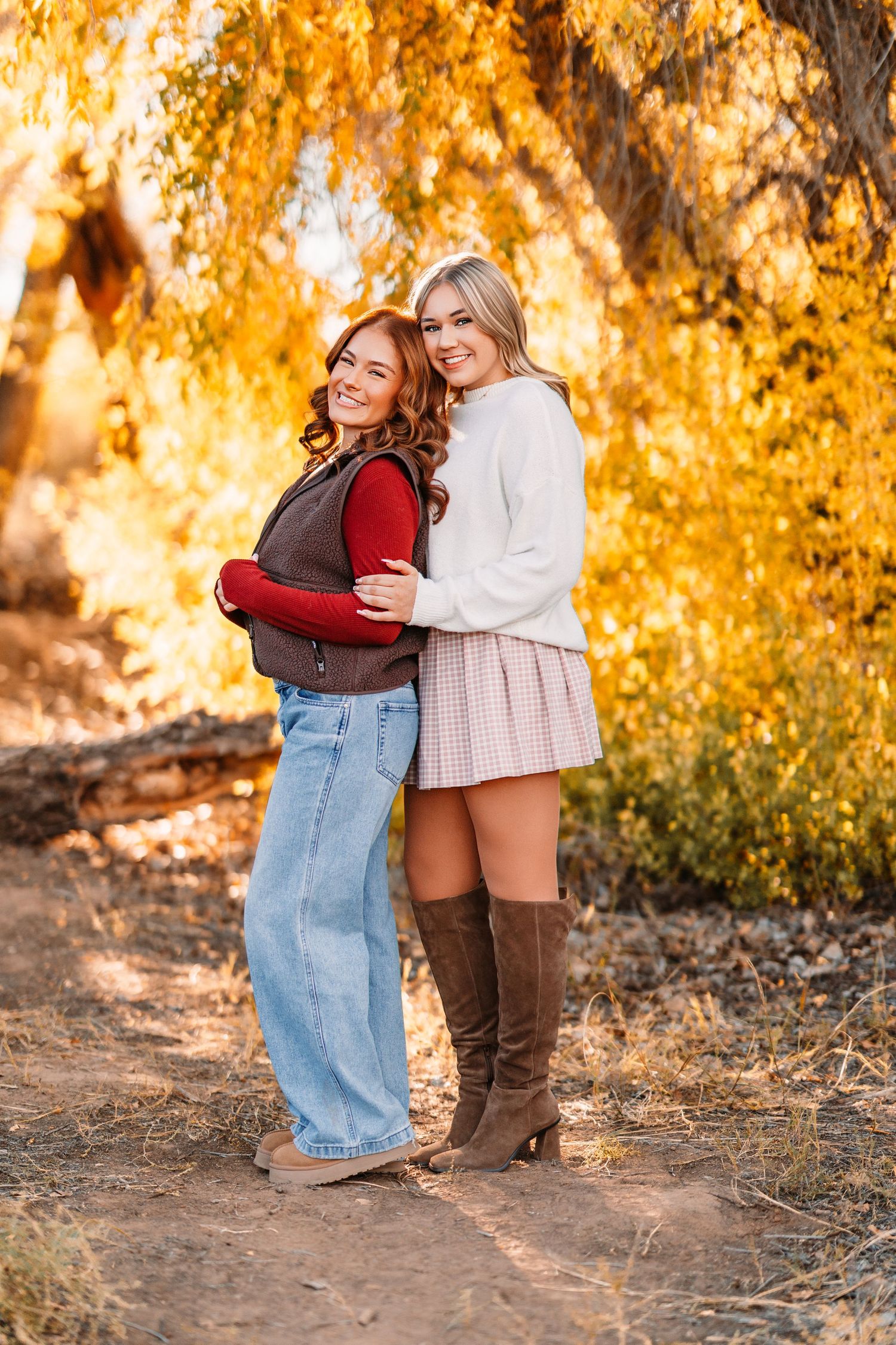 Two friends embrace while wearing fall outfits against a backdrop of golden autumn foliage.