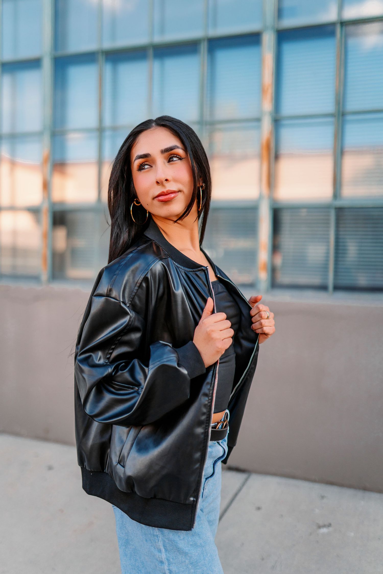 Fashion model in leather jacket and jeans poses against modern glass building in urban photoshoot.