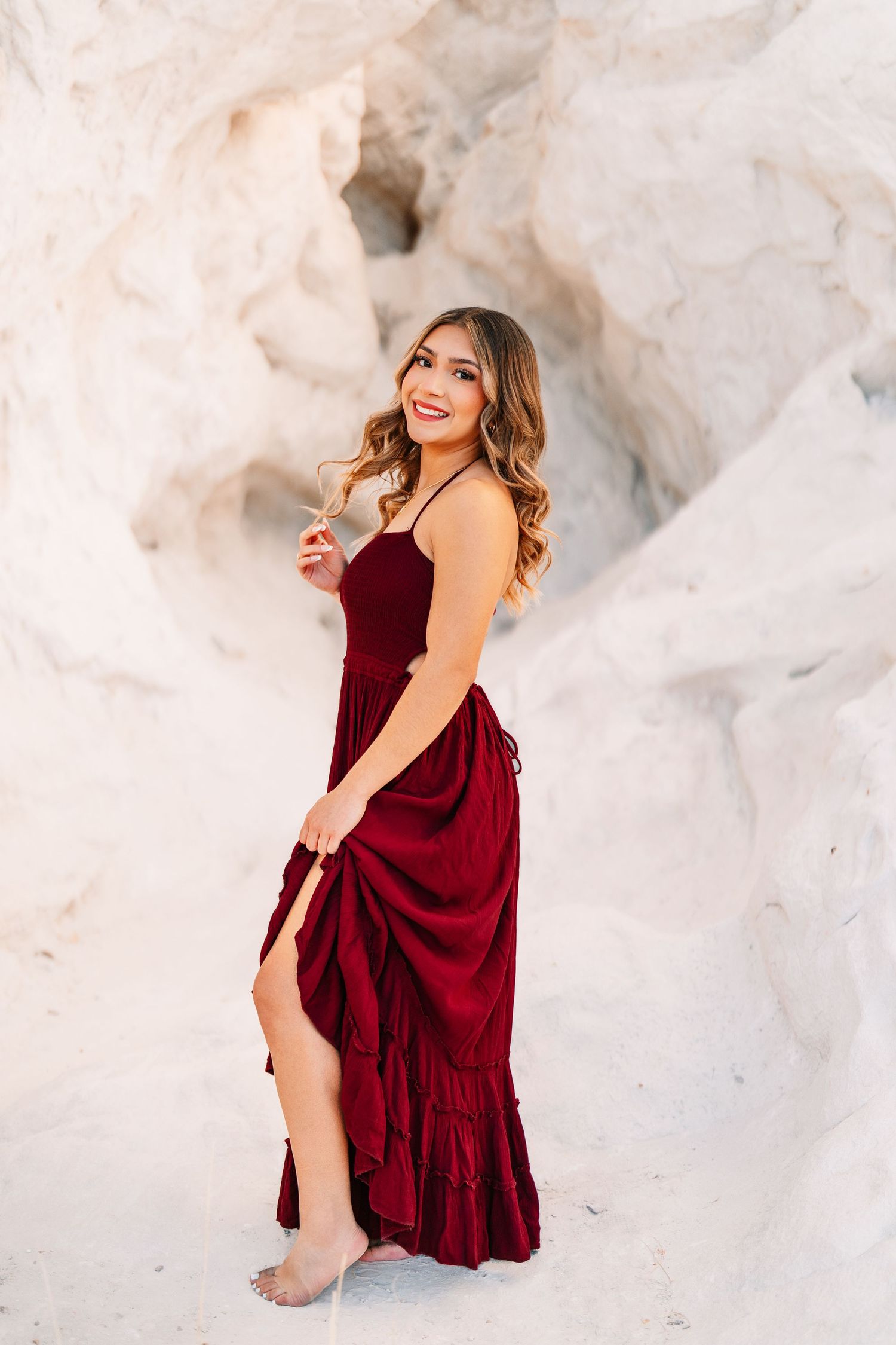 Woman in burgundy dress poses gracefully against white rock formations in outdoor photoshoot.