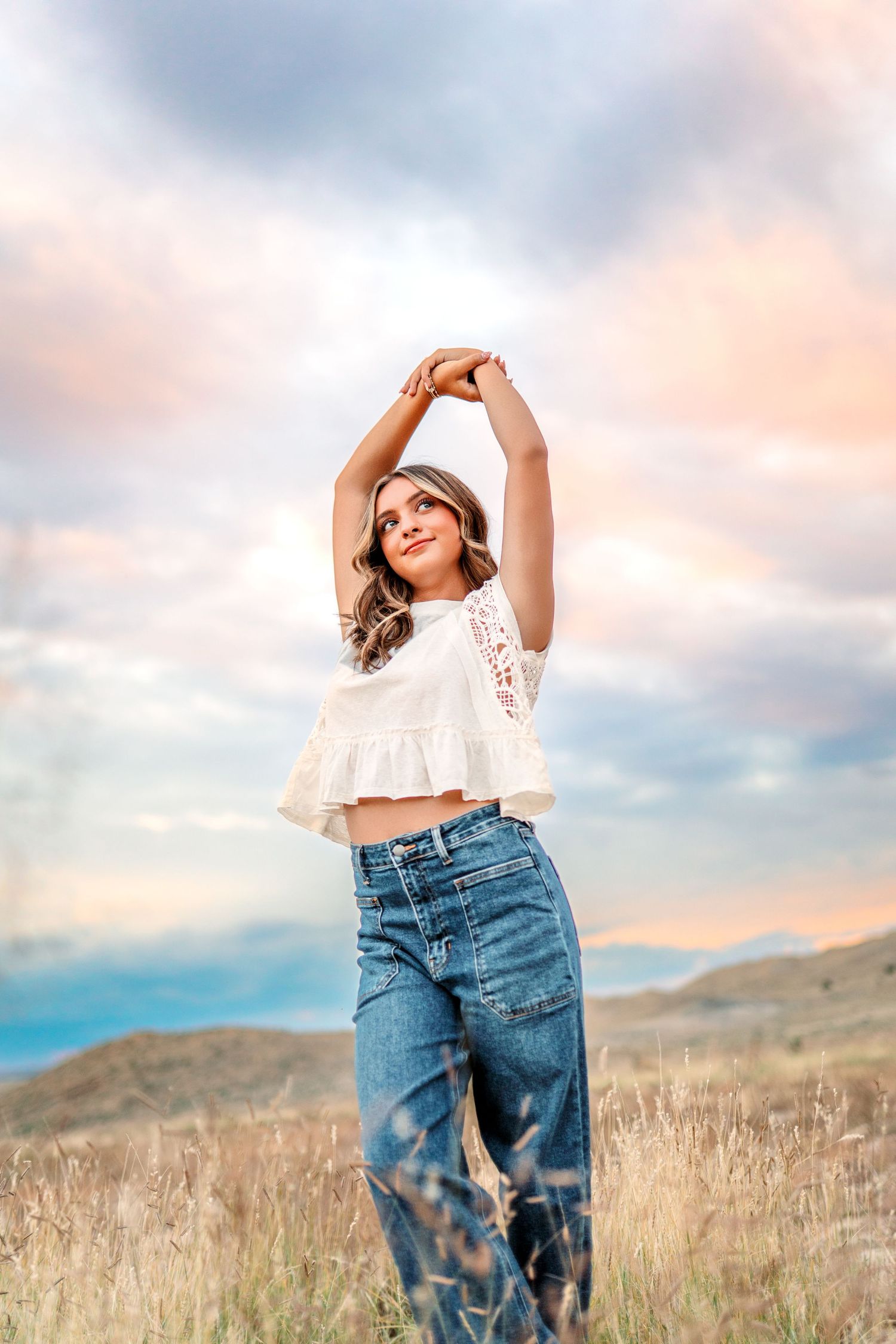 Woman in white crop top and jeans stretches joyfully in field at sunset.
