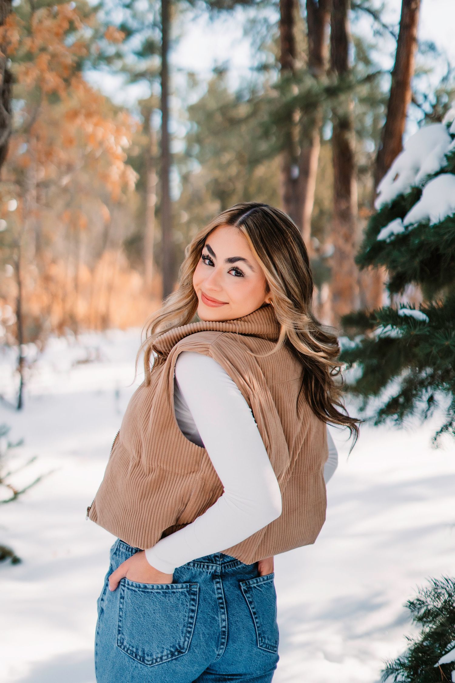 Winter portrait session in a snowy forest wearing tan sweater and blue jeans.