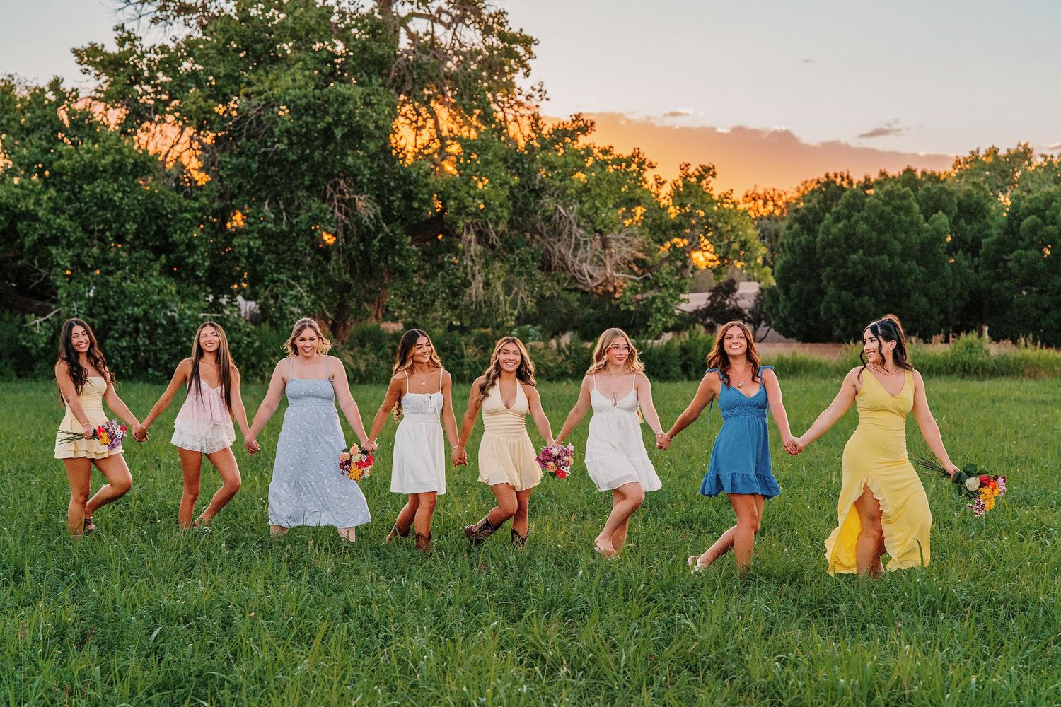 Group holds hands while walking through green field during golden sunset.