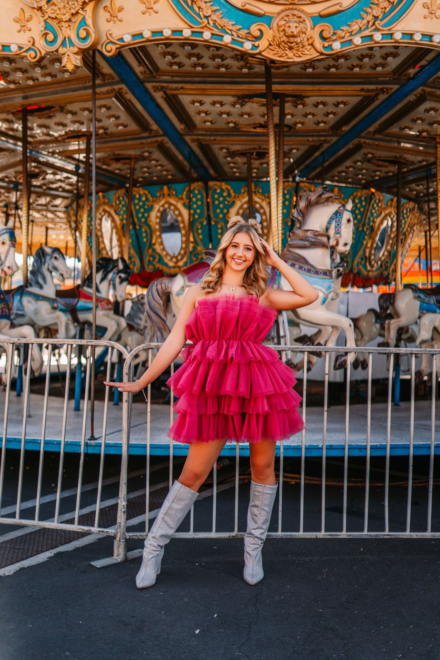 Playful fashion photo at a carnival carousel featuring a hot pink tulle dress and white boots against colorful fairground lights.