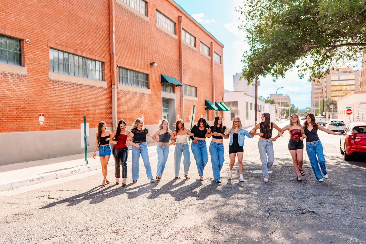 Line of people in casual outfits pose along brick wall with long shadows cast by sunlight.