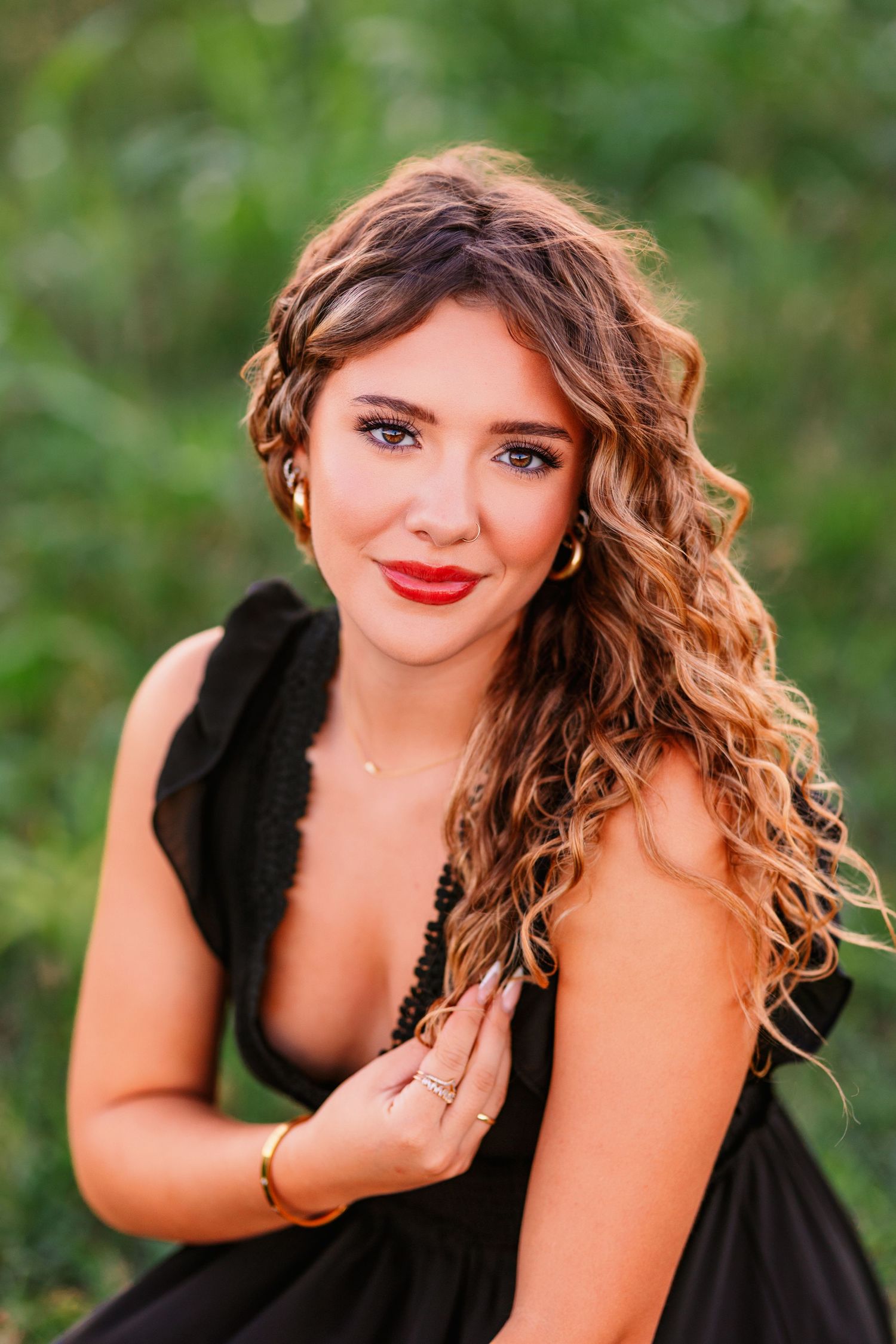 Portrait of someone in a black dress with long wavy brown hair and red lipstick against a blurred green background.