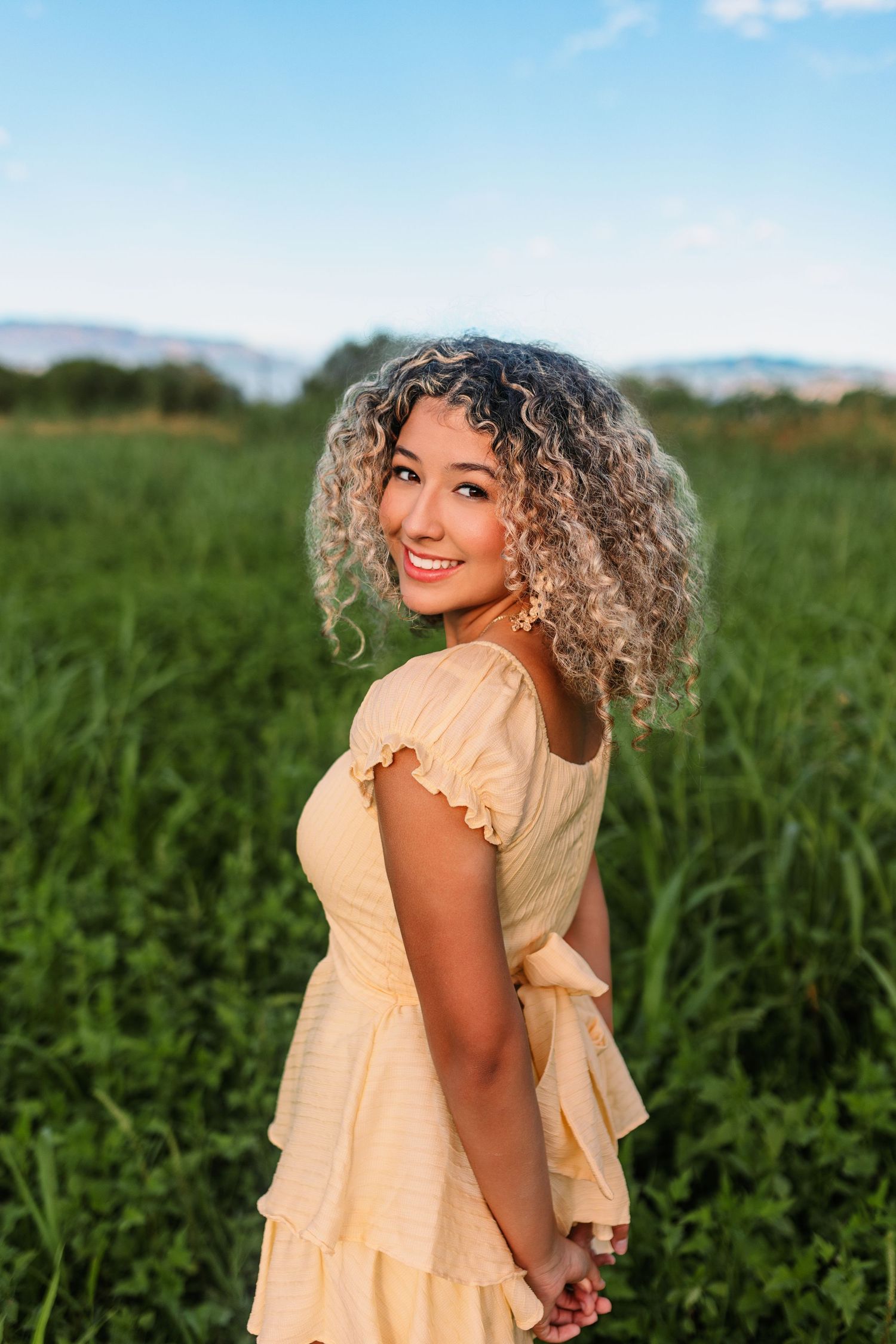A woman in a yellow sundress smiles while standing in a green field during golden hour.
