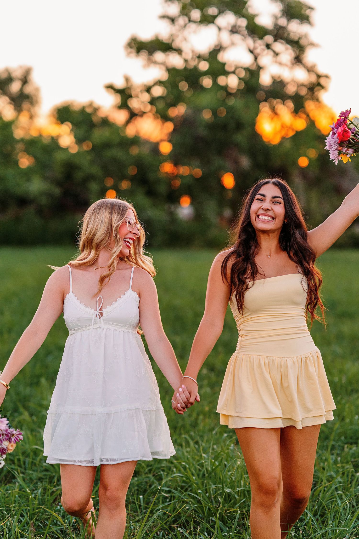 Friends in summer dresses hold hands while walking through a grassy field at sunset.