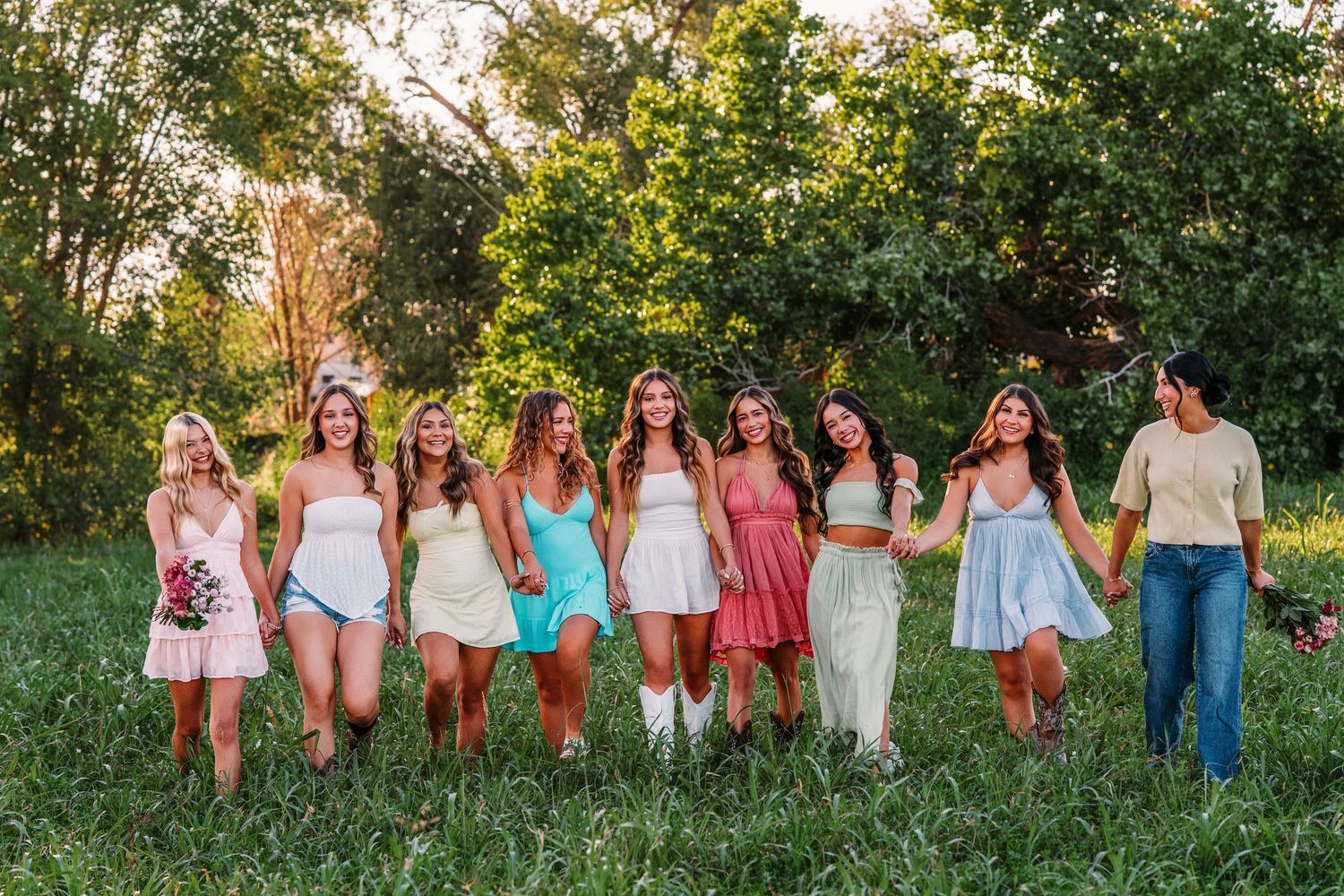Group portrait of friends in summer dresses standing together in a grassy field at sunset.