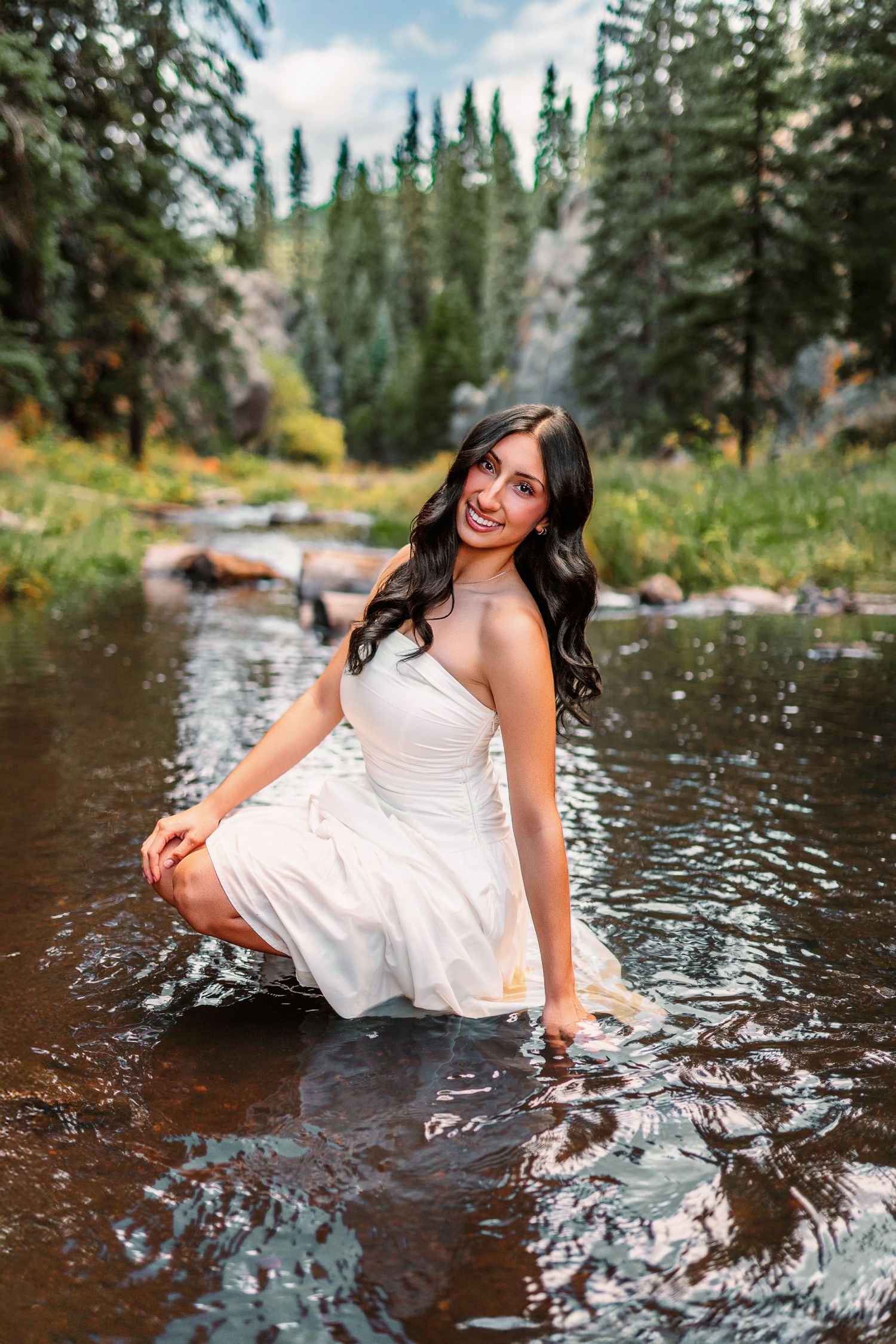 Playful water portraits in white dress at mountain stream with evergreen forest backdrop.