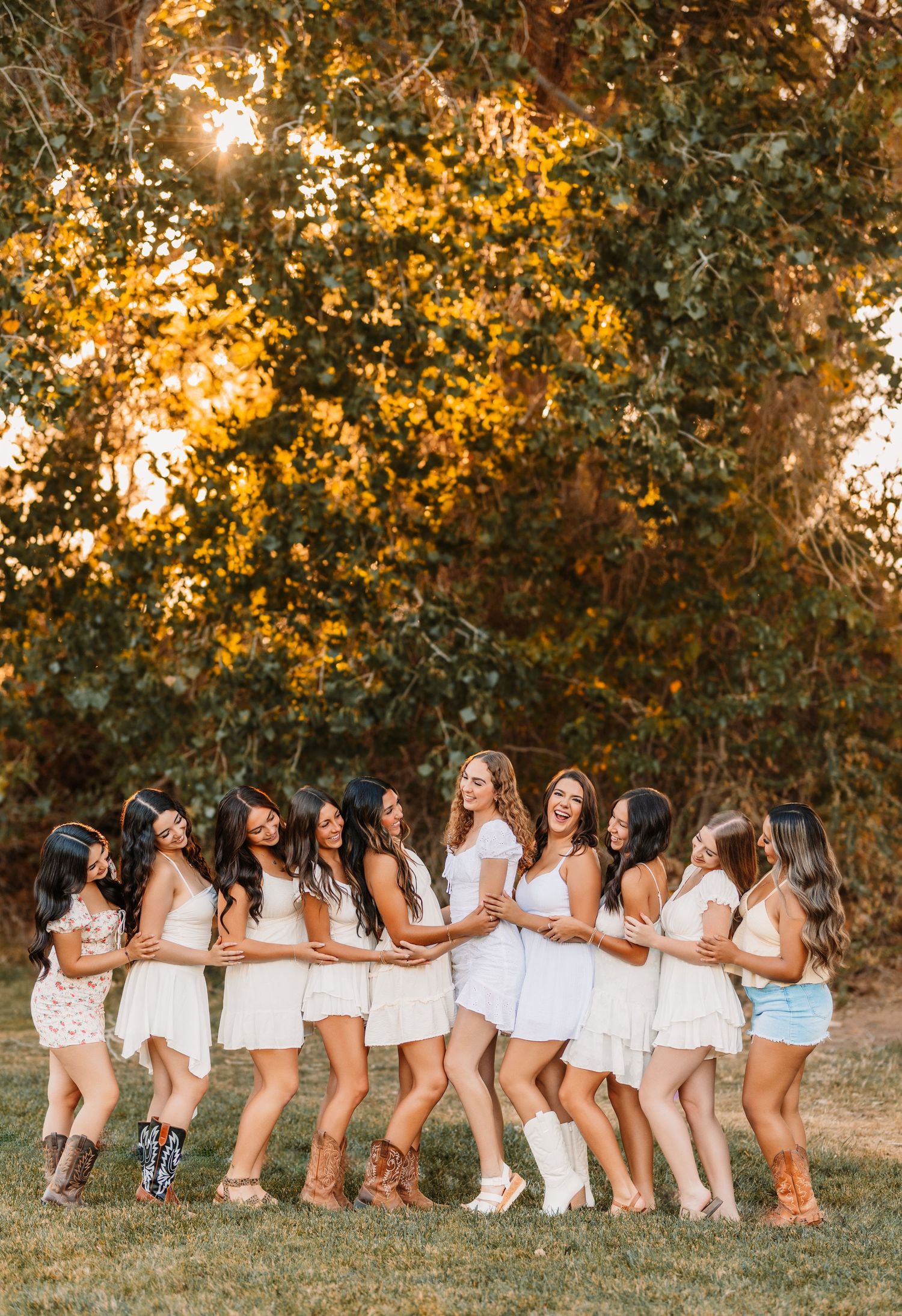 Row of friends in white tops and boots pose together on grass during golden hour.