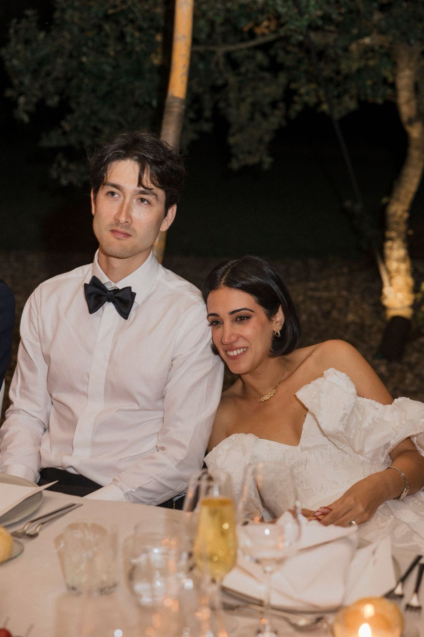 A couple sits together at an elegant outdoor wedding reception table with candlelight and drinks at dusk.