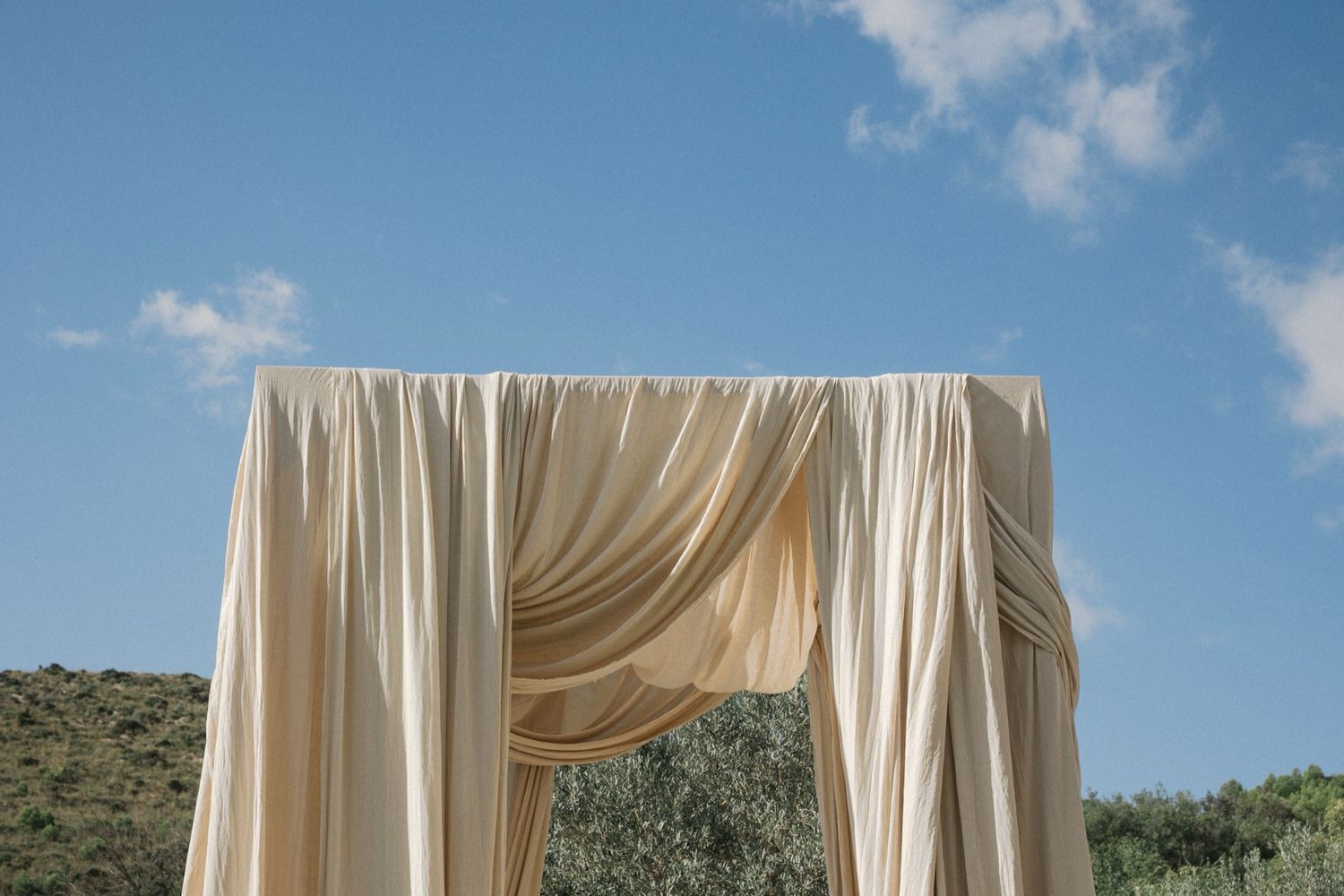 Elegant cream-colored fabric wedding arch draped against blue sky and desert mountain landscape.